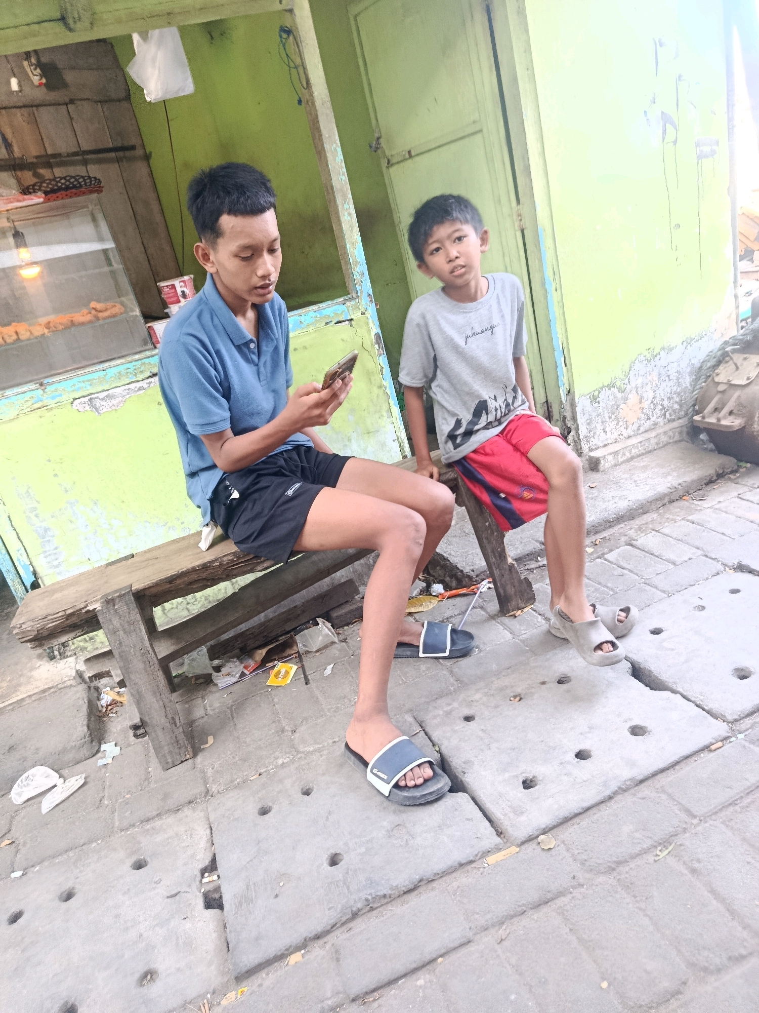 Two young males are seated on a wooden bench outside a food stall with a green facade in Jobokuto, Indonesia. The male on the left, wearing a blue polo shirt and black shorts, is looking down at a smartphone in his hands. The male on the right, wearing a grey t-shirt and red shorts, is looking forward. The food stall appears to be open, with a display case visible behind the male on the left, showing fried food items. The ground is paved with concrete slabs and there is some scattered debris. The overall scene suggests a casual, everyday moment outside a local eatery.