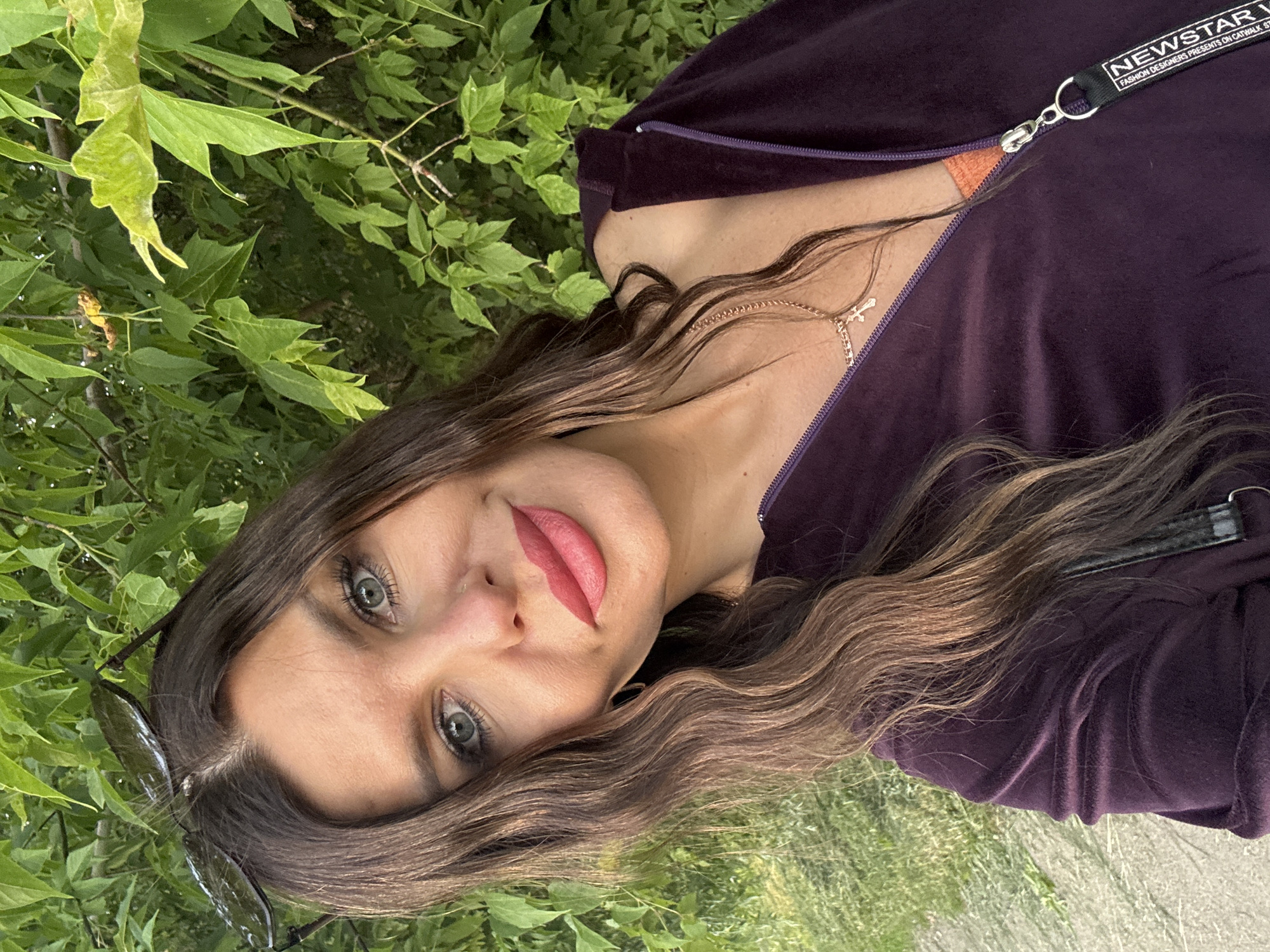 This media file features a close-up, horizontal shot of a woman lying outdoors amidst greenery, possibly in Chaikovsky, Russia. The woman has long, wavy brown hair and striking green eyes. She is wearing a dark purple velour hooded jacket, partially unzipped, revealing an orange top underneath. A delicate gold chain with a cross pendant is visible around her neck. Sunglasses are resting on her cheek, and her lips are painted a vibrant pink. The background consists of lush green leaves and grass, suggesting an outdoor setting during daytime with fair weather. The overall mood is relaxed and natural. Visible text on a black tag attached to the zipper reads "NEWSTAR" followed by smaller text that appears to be "FASHION DESIGNERS PRESENTS ON CATWALK ST".
