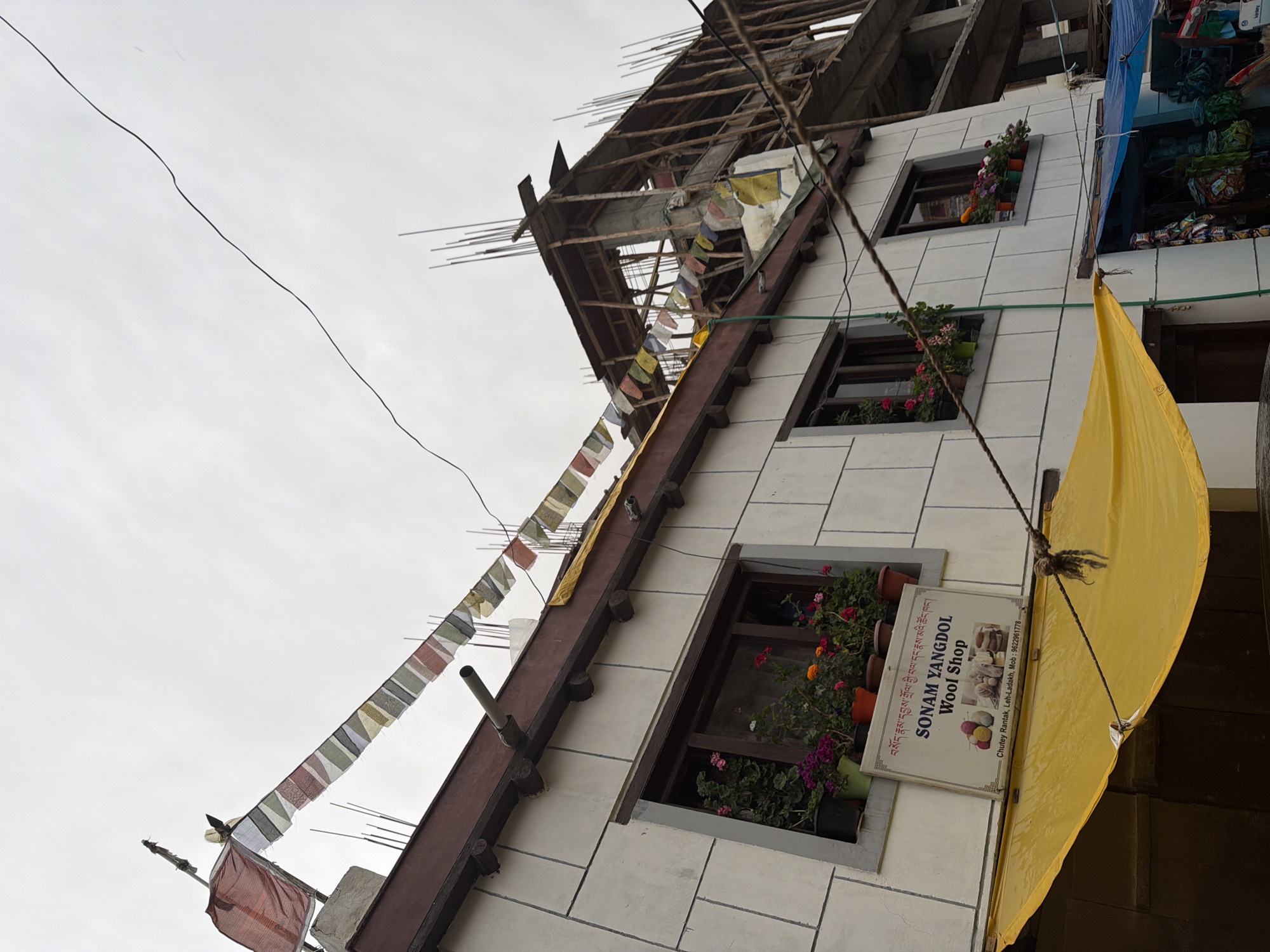 The image depicts the exterior of a building in Leh, India, with a cloudy sky overhead. Prayer flags are strung across the front of the building, partially obscuring a construction site on the right. Below the flags, there are two windows with flower boxes filled with blooming plants. A sign beneath the right window reads "SONAM YANGDOL WOOL SHOP," with a phone number listed as "Mob: 9622961778" and an address "Chutey Rantak, Leh-Ladakh". A bright yellow tarp is stretched across the lower portion of the building, covering an entrance. To the right of the yellow tarp, a blue tarp covers what appears to be a market stall with various items displayed. The building itself has a white facade with grey grout lines. The construction site on the right shows exposed concrete and wooden scaffolding.