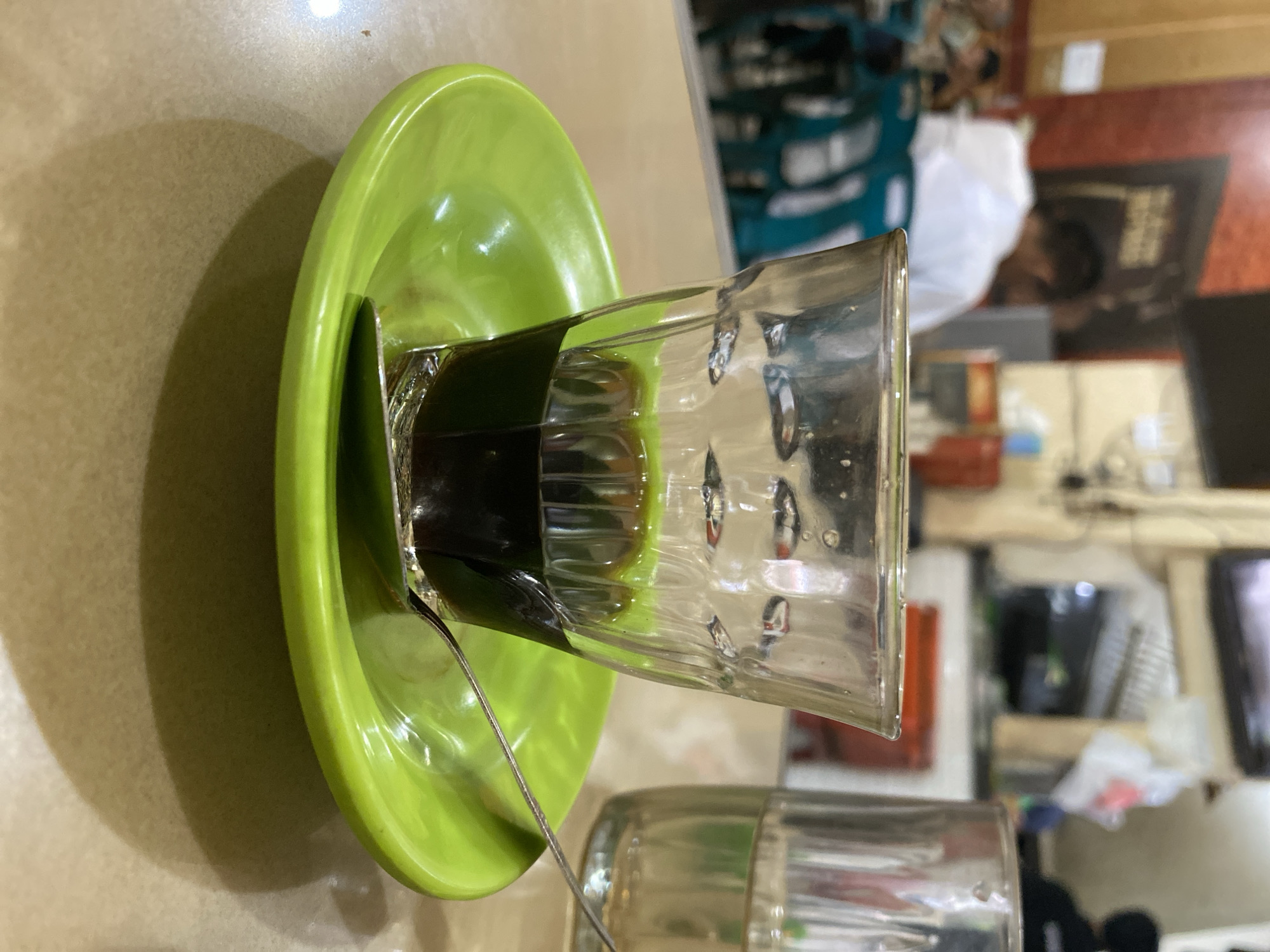 The image shows a close-up of a glass of dark liquid, likely coffee, with a greenish hue, sitting on a bright green saucer. A silver spoon rests on the saucer. In the background, out of focus, is a dimly lit indoor setting that appears to be a cafe or restaurant. Several people are visible, some seated at tables, and a counter area with what might be a refrigerator or display case. The overall ambiance suggests a casual, public eating establishment. Based on the visual context and the user's prompt, this scene is likely located in a cafe in Lhokseumawe, Indonesia.