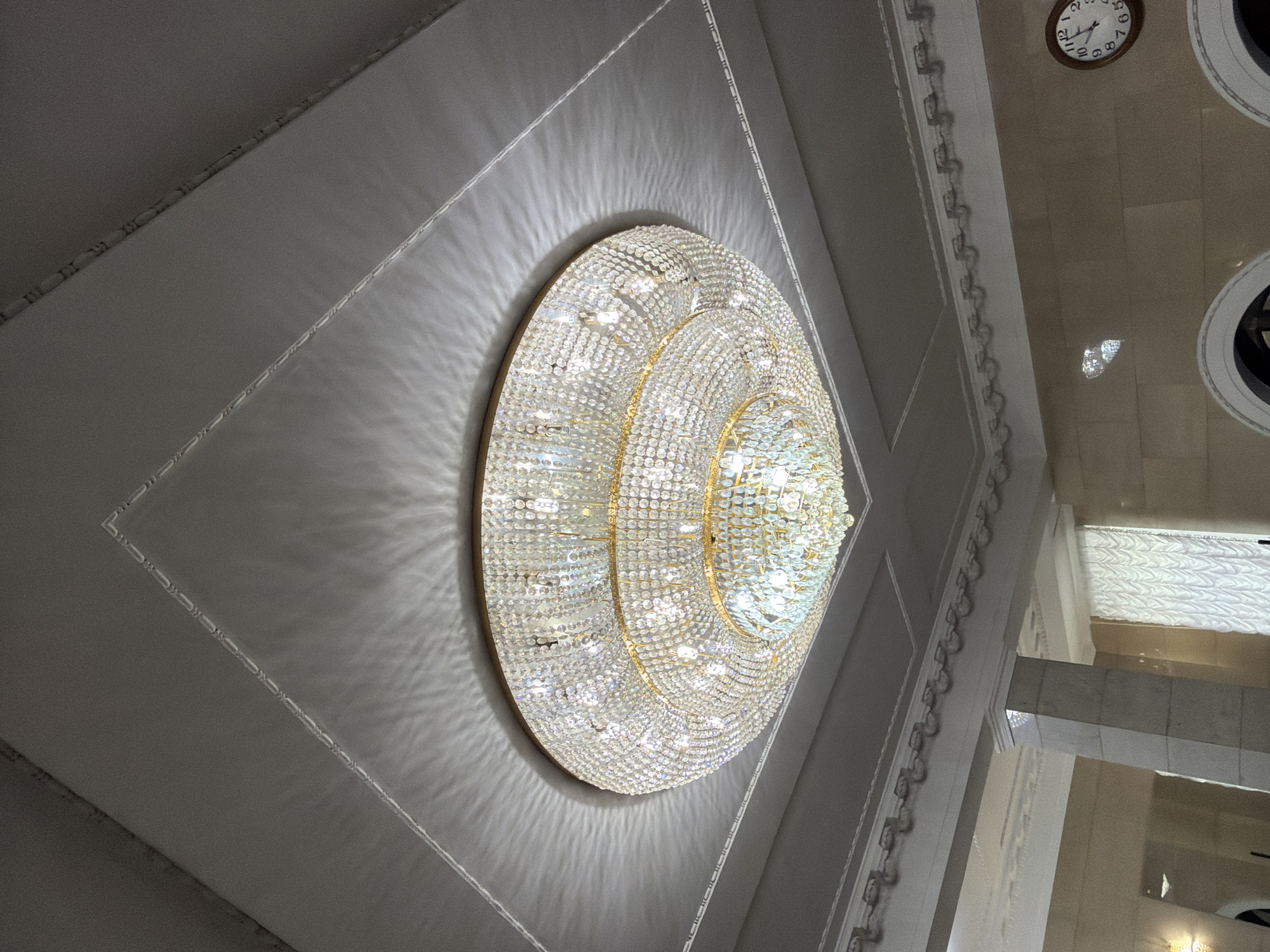 A large, multi-tiered crystal chandelier hangs from a white ceiling in a room. The ceiling has recessed square paneling with ornate trim. The chandelier is round and consists of many crystal droplets and golden accents. Light from the chandelier casts intricate shadows on the ceiling. Below the chandelier, architectural columns and decorative molding are visible. A clock on the wall shows the time as approximately 1:10.