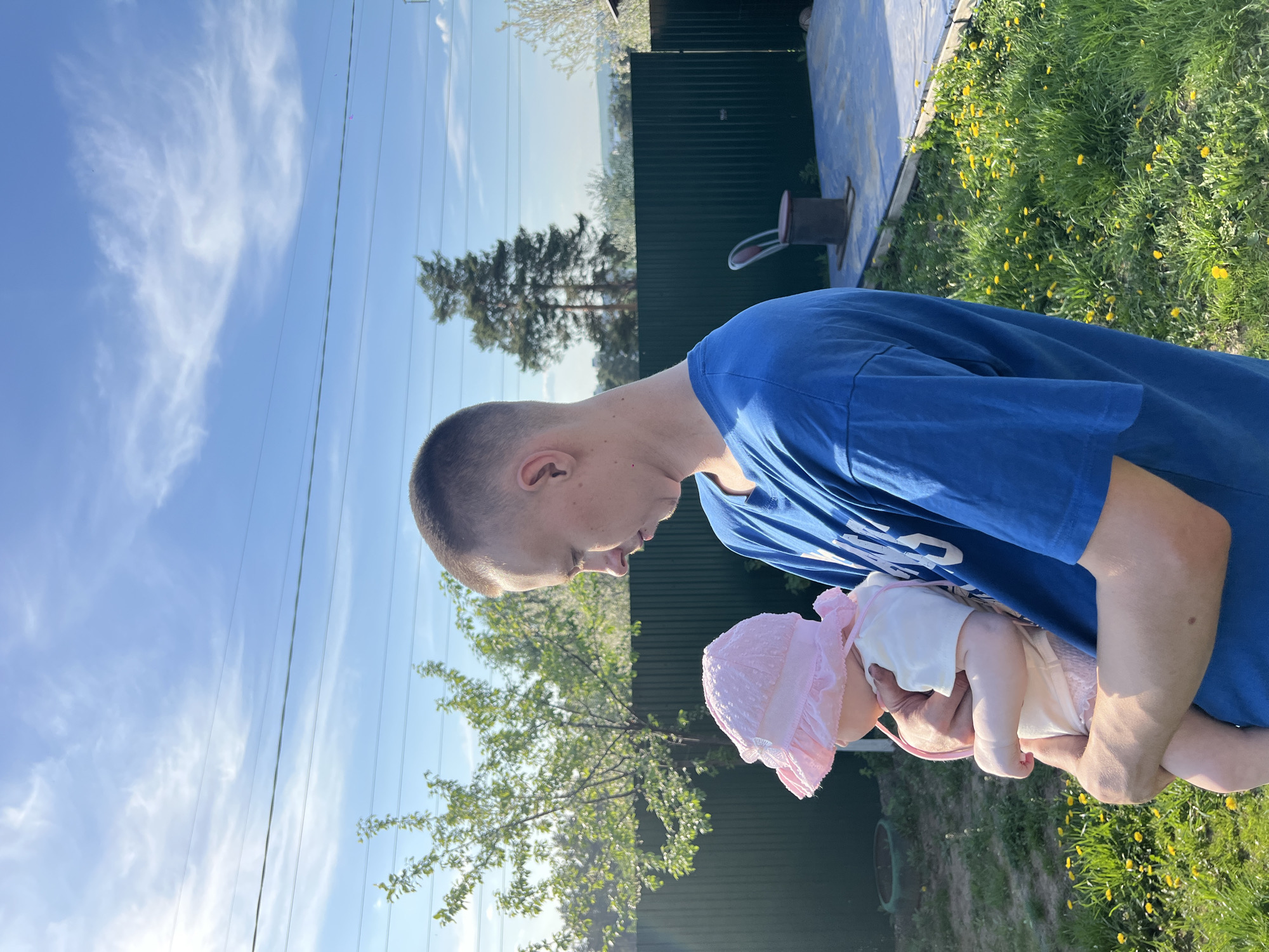 A man in a blue t-shirt is holding a baby with a pink hat in an outdoor setting. The location appears to be in a grassy area, possibly a backyard, near a fence. The sky is visible in the background, suggesting it is daytime. The picture appears to be rotated. No evidence indicates the location is Chaikovsky, Russia.