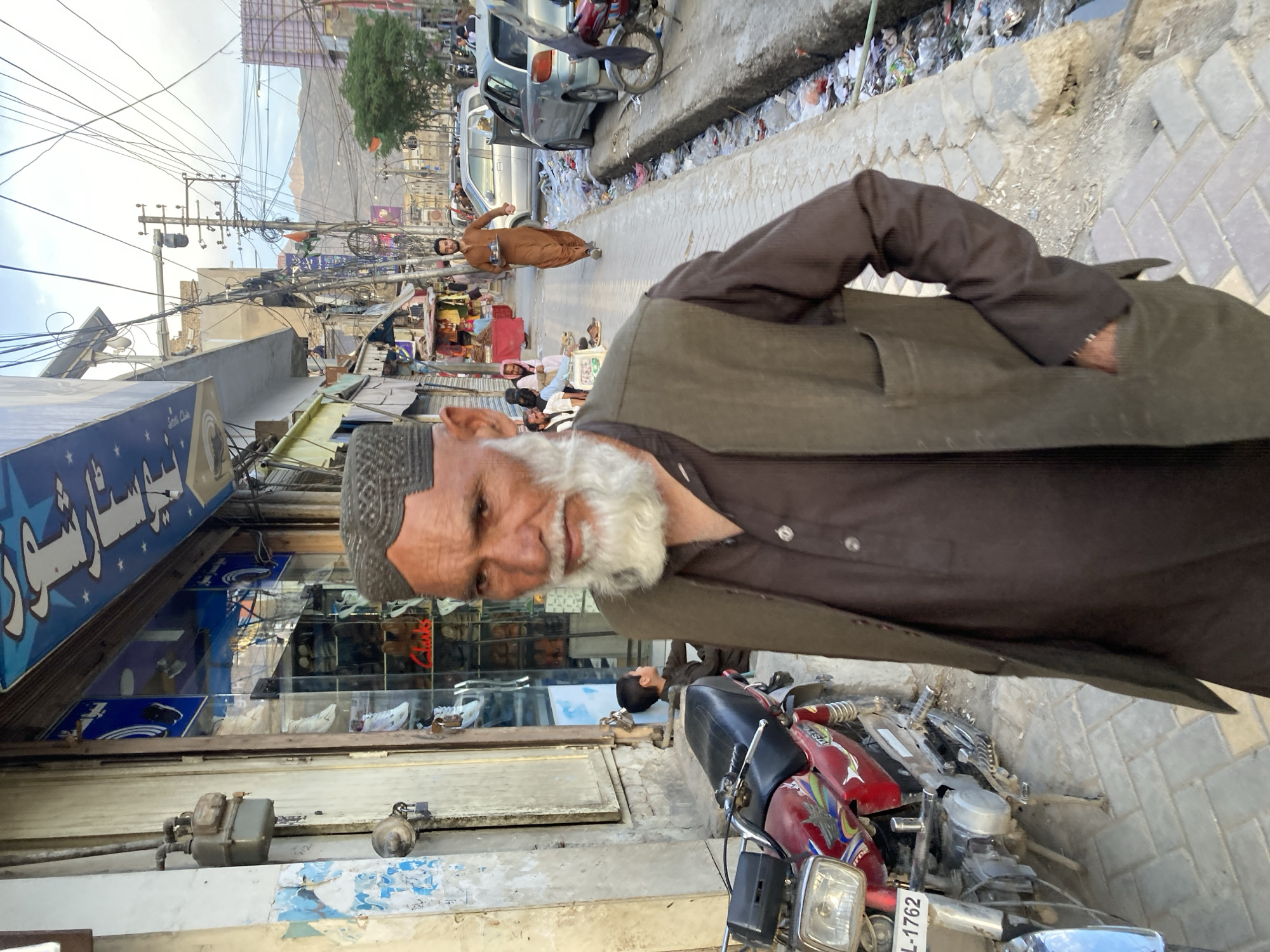 An eye-level photograph in Quetta City Tehsil, Pakistan, shows an elderly man with a white beard, wearing a dark button-down shirt and vest, standing on a street near a parked red and blue motorcycle. The street extends into the background, revealing shops, other people, and vehicles. In the distance, mountains are visible under a cloudy sky. Overhead power lines add to the urban landscape. Debris and trash line the edge of the road.