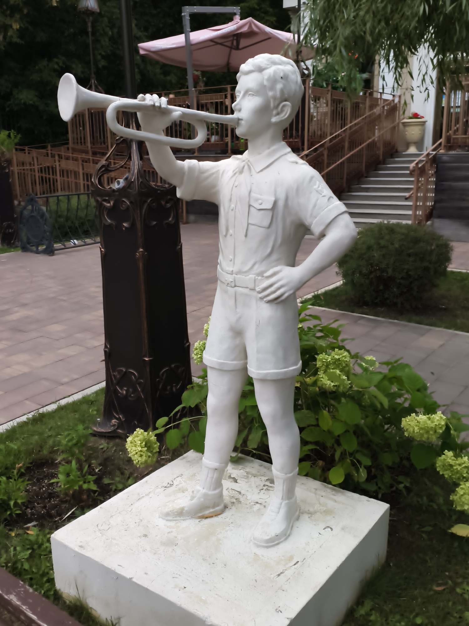 A full-body, white stone statue of a young boy playing a trumpet stands on a white pedestal in a grassy area. The statue depicts the boy in a short-sleeved collared shirt with pockets, shorts, and high socks with shoes. He holds a trumpet with both hands, bringing it to his lips. The boy's left hand is on the trumpet's valves. His right hand rests on his hip. He is facing slightly to the right of the viewer. Behind the statue, a dark ornate metal post stands to the left, and a dense green bush is behind the boy's right leg. In the background, a paved walkway is visible to the left, and stairs with ornate railings lead up to a building or structure. Green foliage and a pink umbrella are also visible in the background. The ground around the statue is covered in grass and some dirt. The lighting suggests it is daytime, possibly overcast.