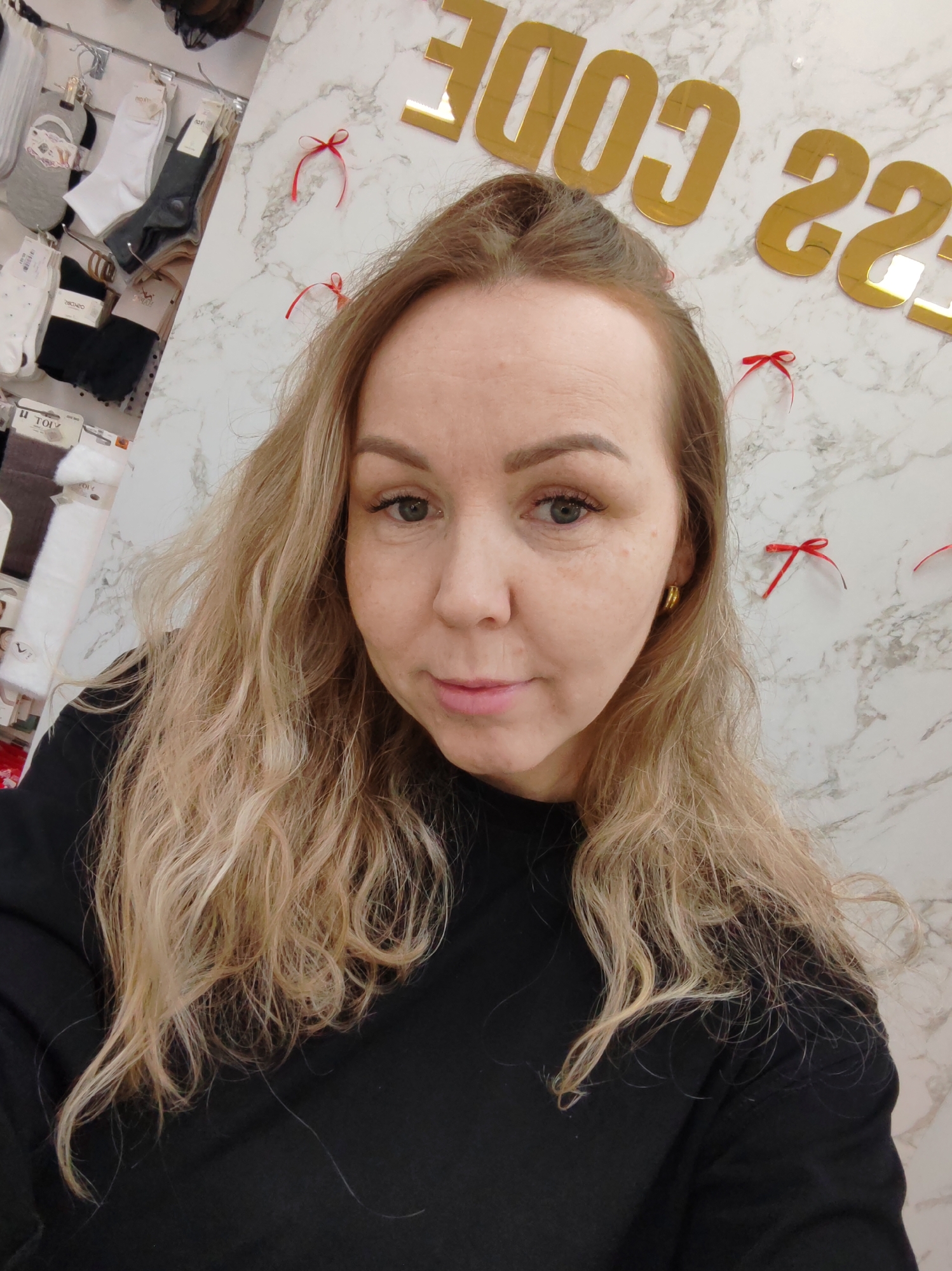 A medium shot captures a blonde woman in Chaikovsky, Russia, indoors, likely in a store or commercial setting. She wears a black long-sleeved shirt. Behind her is a marbled wall adorned with small red bows and gold lettering that reads "DRESS CODE." To the left, socks hang on a display rack. The lighting is bright and even.
