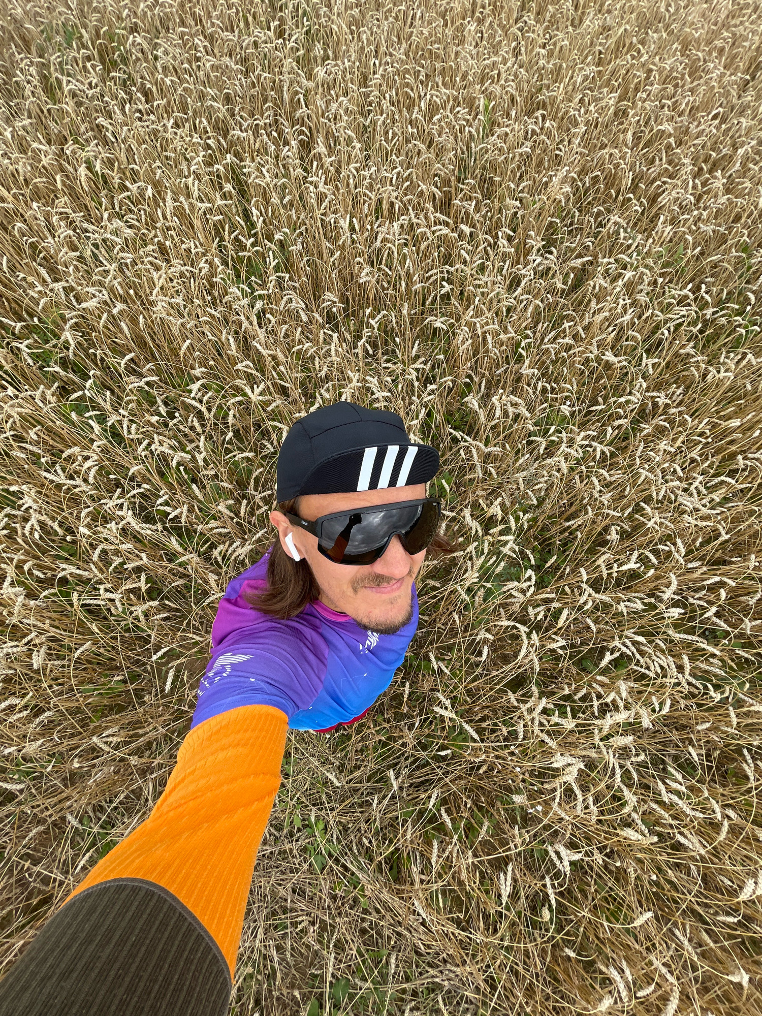 A man is taking a selfie in a field of wheat in Perevoznoe, Russia. He is wearing a black cap with white stripes, sunglasses, white wireless earbuds, and a colorful athletic shirt with purple, blue, and orange accents. His right arm, extended towards the camera, is covered in an orange sleeve. The field of wheat is dry and golden, covering the entire background.