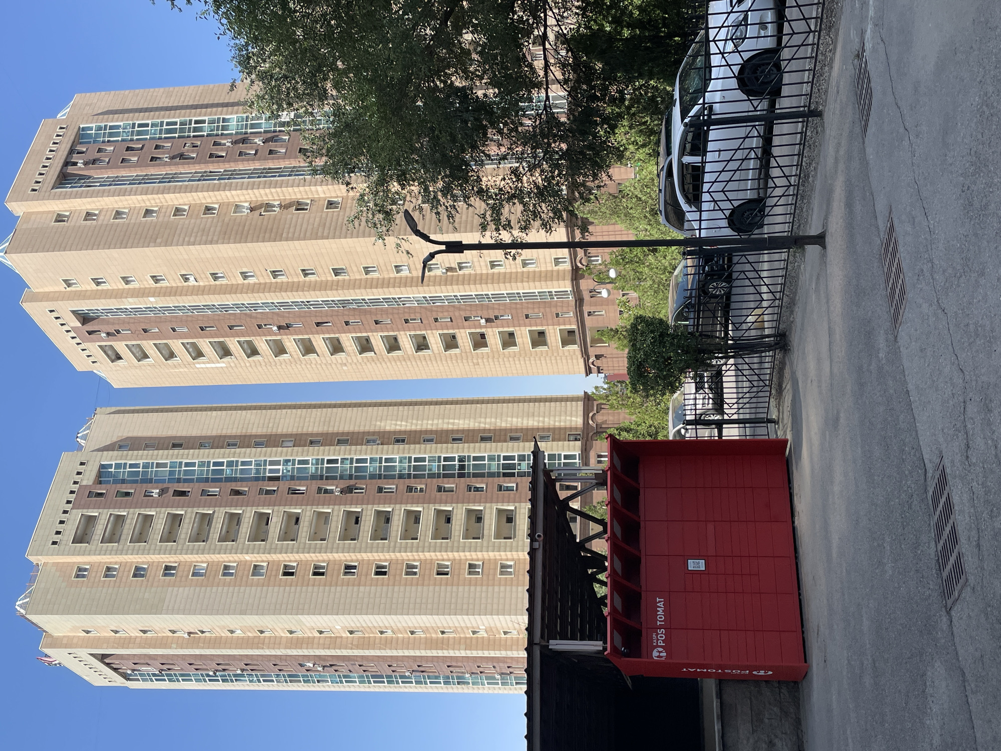 The image depicts two modern, beige and brown high-rise buildings in Almaty, Kazakhstan. The buildings have many floors with balconies and large glass windows. In the foreground, a bright red parcel locker station labeled "KASPI POS TOMAT" is visible. Next to it, a black metal fence encloses a parking area where several cars are parked, including a white sedan. The environment is an urban setting with a clear blue sky and some trees.