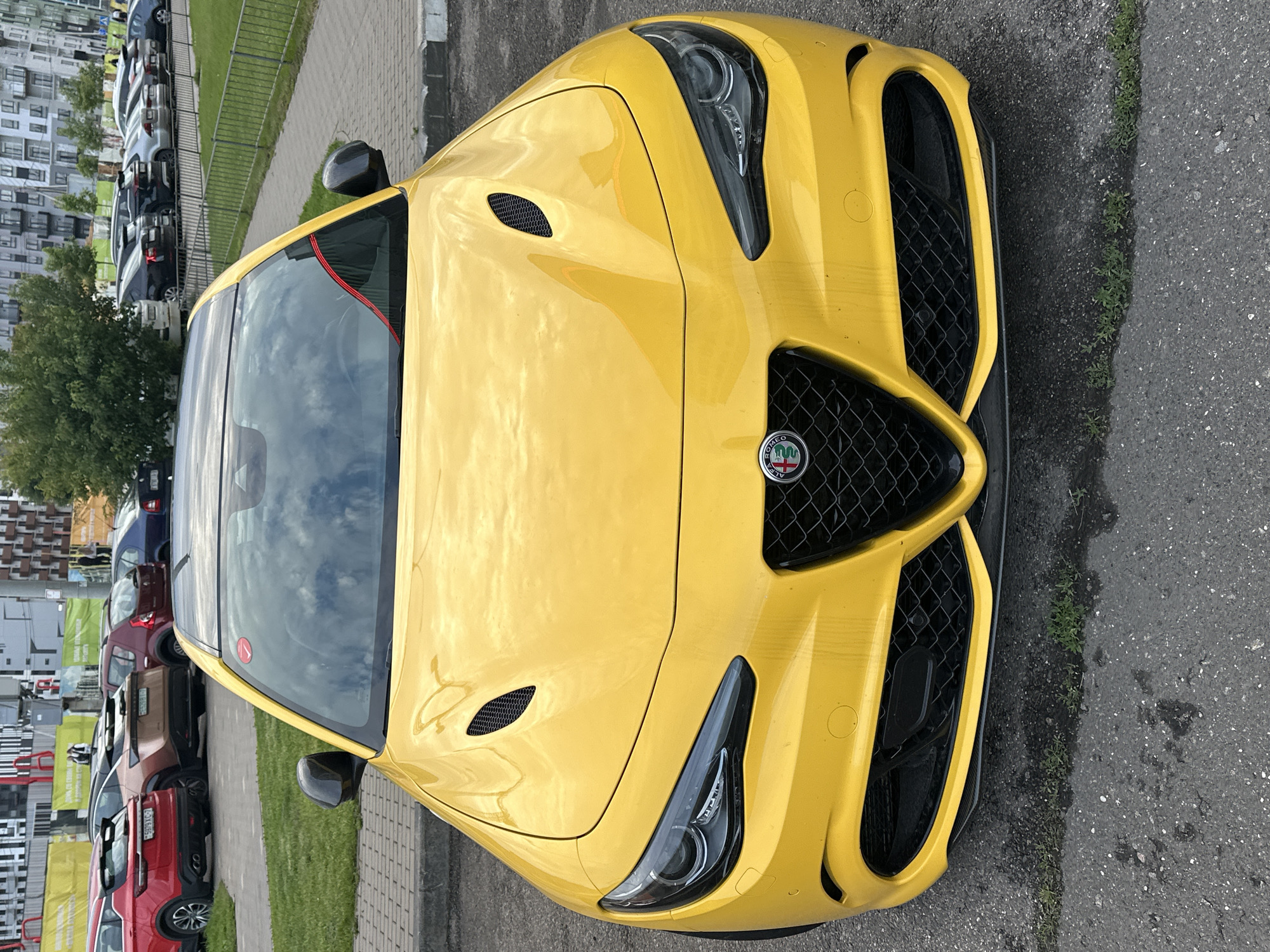 This is a vertical, eye-level, close-up photograph of the front of a bright yellow sports car. The car's hood and front bumper dominate the frame, with the grille and headlights prominently featured. The car has aggressive styling, with sharp lines and large air intakes. The grille has a honeycomb pattern and the Alfa Romeo logo is visible in the center. The headlights are sleek and modern.
In the background, to the left of the car, there is a line of parked cars and buildings, suggesting an urban or suburban setting. There is also a patch of green grass and a sidewalk. The sky, reflected in the car's windshield, is cloudy. The lighting suggests it could be daytime, possibly overcast. The image appears to be taken outdoors. No people are visible in the photograph. There is no visible text in the image. The car appears to be stationary.