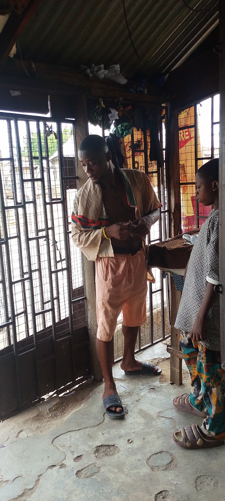 The image depicts two individuals standing in what appears to be a makeshift dwelling in Lokoja, Nigeria. The first individual, a young man, is fair-skinned and of average height. He is wearing peach-colored shorts and a tan shirt with red and green stripes. He is also wearing dark blue plastic sandals. He has a bracelet on his left wrist and appears to be looking down, possibly at his hands. His torso is exposed. The second individual, a child, is standing to the right, partially obscured by the doorway. They are wearing a patterned white and black shirt and pants with a colorful, abstract pattern. They are wearing brown plastic sandals. The structure they are in has a corrugated metal roof, a barred window, and a partial wall. The floor is concrete with visible wear and damage. There are clothes and other items stored above the doorway. The scene appears to be taken outdoors, in daylight.
