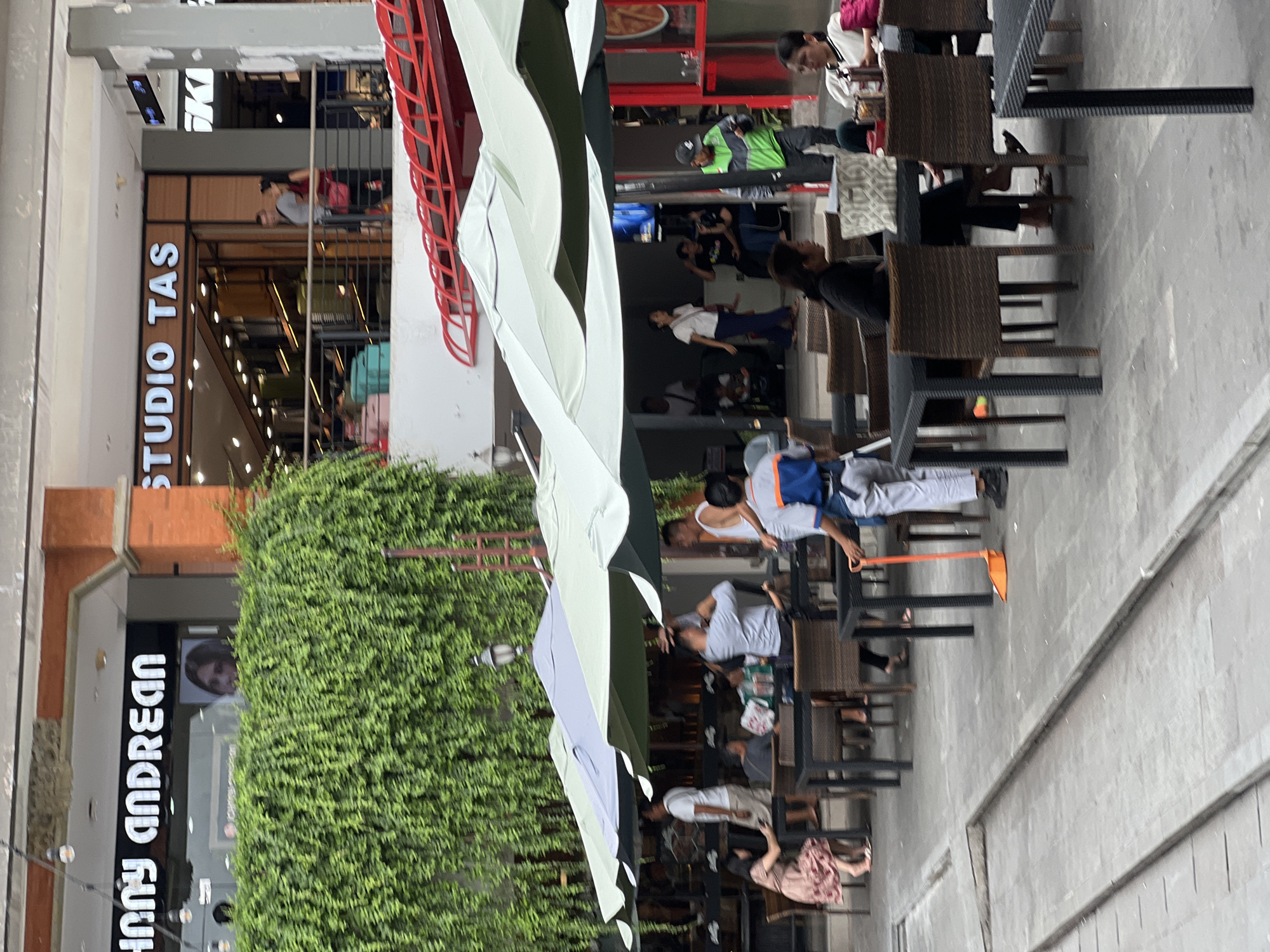 The image depicts a street scene in Kuta, Indonesia. The shot appears to be taken near a building with storefronts including "STUDIO TAS" and "JOHNNY ANDREAN". Pedestrians are visible, some sitting at outdoor tables and chairs near the stores, others are walking nearby. A maintenance worker sweeps the street with a broom. The area is partially shaded by canopies of some kind. A significant amount of greenery, likely a large hedge or wall of plants, is also visible. The environment has an open, public feel to it.