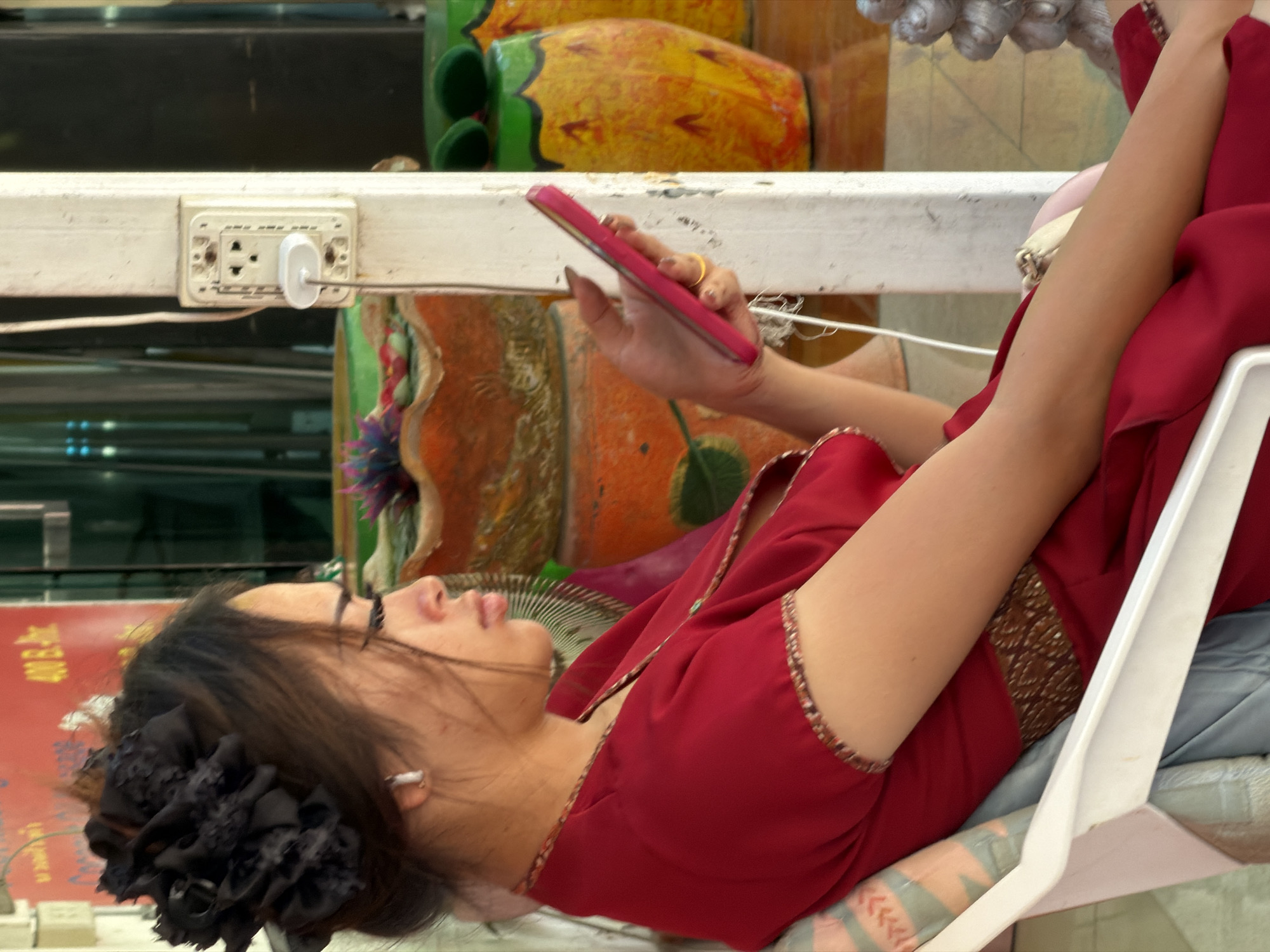 A young woman with dark hair adorned with a large black fabric flower and a small silver earring is seated on a white plastic chair, facing right in profile. She is wearing a red dress with a gold-toned patterned trim at the neckline and sleeves. The woman is holding a pink smartphone in her right hand, which is resting on her lap. Her left arm is bent, with her elbow resting on the armrest of the chair. In the background, a white wooden post with an electrical outlet is visible. Behind the post, there are decorative potted plants, one orange and patterned, and others resembling pineapples. The setting appears to be an outdoor area, likely a street or a market stall, in the Ko Samui District of Thailand, judging by the presence of the electrical outlet and the style of the decorative items.
