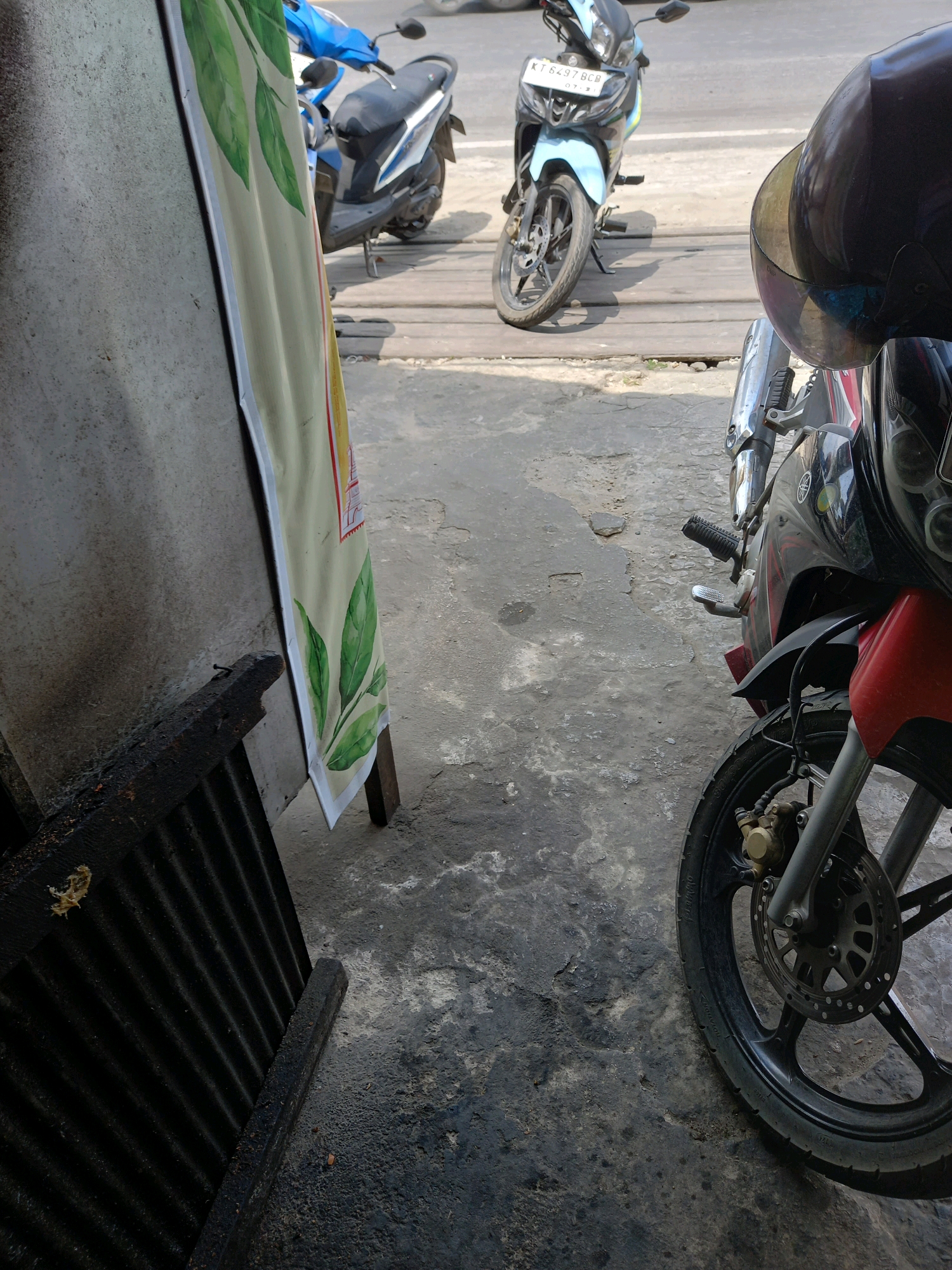 The image depicts an outdoor scene in a city, likely in Samarinda, Indonesia, judging by the visible license plate which reads "KT 6497 BCB". In the foreground, a black and red motorcycle is parked, with its front wheel and braking system prominently displayed. The motorcycle has a red front fender and a black body with red accents. A portion of a black helmet is visible in the upper right corner of the frame, suggesting a person was recently present or is about to operate the motorcycle.
Behind the foreground motorcycle, a blue and white motorcycle is parked. This motorcycle has a license plate indicating it is registered in Kalimantan Timur. Further in the background, another motorcycle, predominantly light blue, is also parked. The motorcycles are positioned on a surface that appears to be a mix of concrete and wooden planks, suggesting a road or walkway. A large, light-colored banner with green leaf illustrations hangs vertically on the left side of the image, leaning against a textured, grey wall. The overall environment suggests an urban setting, possibly a parking area or a street side.