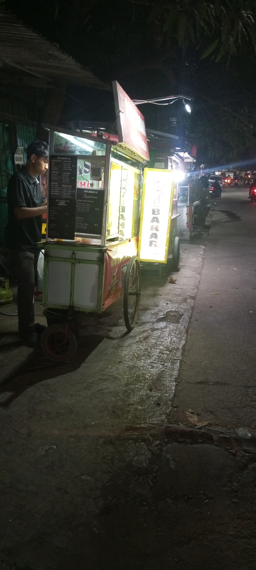 At night, a man in a black polo shirt and dark pants stands next to a food cart on a street in Setu, Indonesia. The food cart is illuminated by bright yellow signs and a white light above. One sign on the cart reads "ROTI BAKAR AJ." The cart is adorned with red and green accents and has two large wheels at the rear and a smaller wheel at the front. A gas tank is visible to the left of the cart. The street surface is wet and reflective, suggesting recent rain. Trees with foliage are visible in the background, and the street is lined with other structures and illuminated by streetlights. Several motorcycles are seen on the road in the distance, their headlights creating streaks of light.