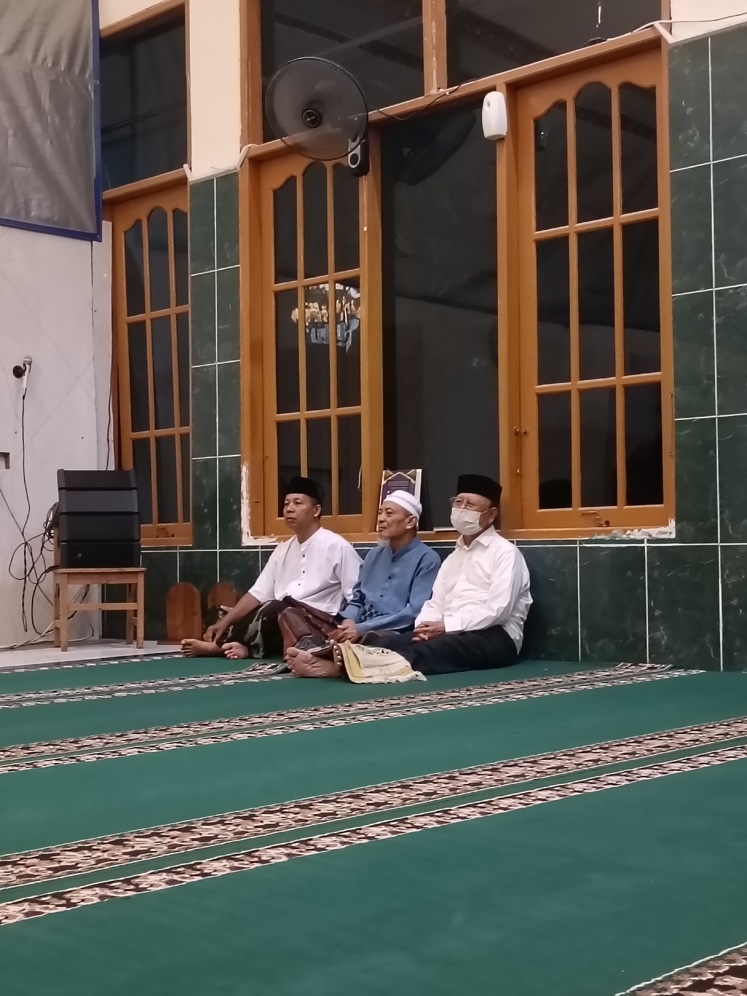 Three men are seated on a green carpet with patterned borders in what appears to be a prayer room or mosque. The man on the left wears a white long-sleeved shirt and a black cap, with a patterned sarong. He is looking to his left. The middle man wears a blue long-sleeved shirt and a white skullcap, and he is also looking to his left. The man on the right wears a white long-sleeved shirt, a black cap, and a face mask. He is looking forward. Behind the men, there are wooden-framed windows with multiple panes and dark interior glass. The walls are tiled in a dark green color with white grout lines. A black speaker system is stacked on a small wooden stool to the left of the frame. A ceiling fan is visible in the upper left quadrant of the image. The setting suggests a cultural or religious gathering in Kediri City, Indonesia.