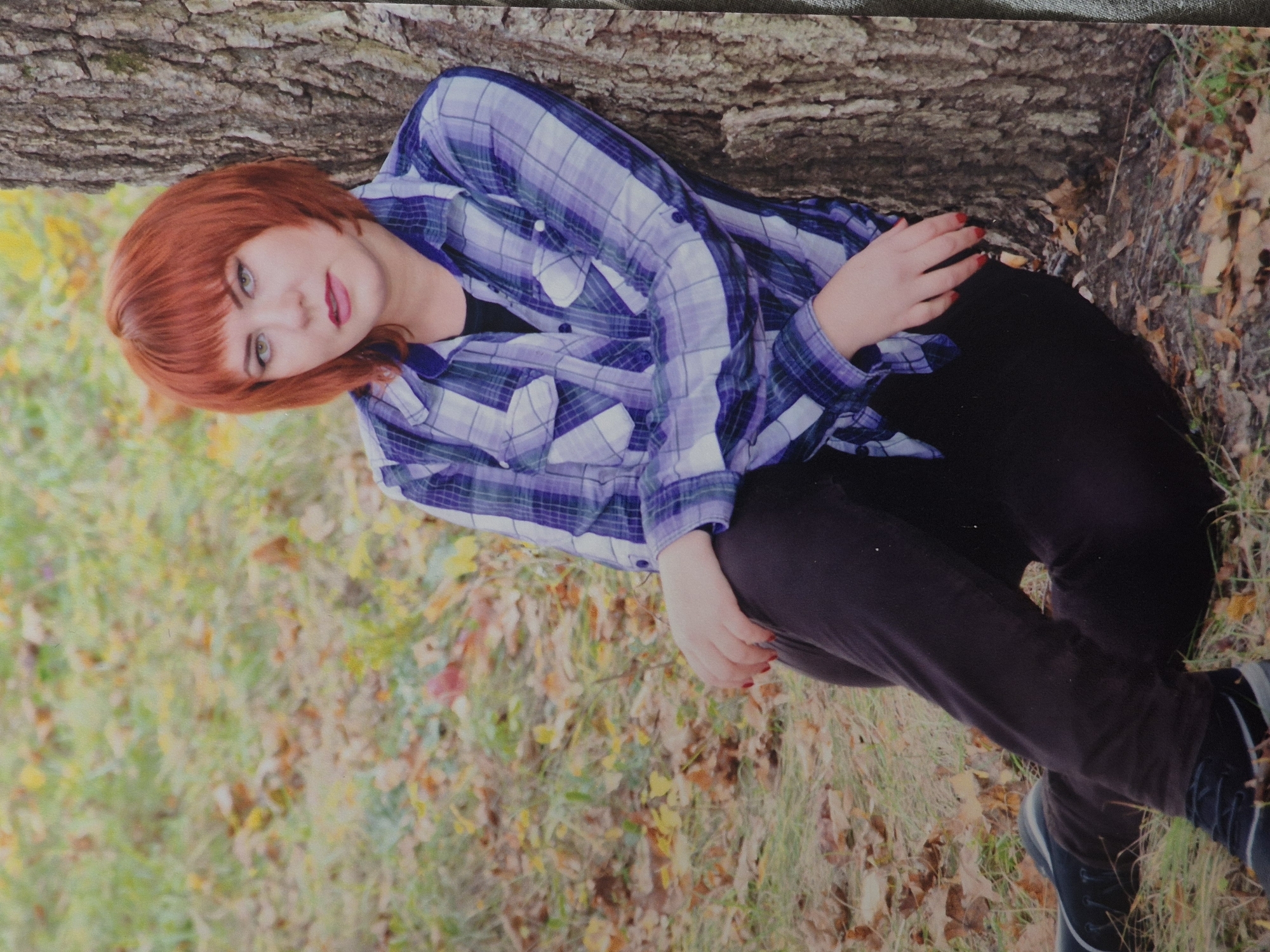 In Schwäbisch Gmünd, Germany, a young woman with short, reddish-orange hair and dark eyeliner sits leaning against a large tree trunk in an outdoor setting. She is wearing a blue and purple plaid button-down shirt over a black undershirt, and dark pants. Her legs are crossed, and her left hand rests on her left knee. Her fingernails are painted red. The ground around her is covered with dry leaves and grass, suggesting an autumn season. The background is a blurred mix of green and yellow foliage.