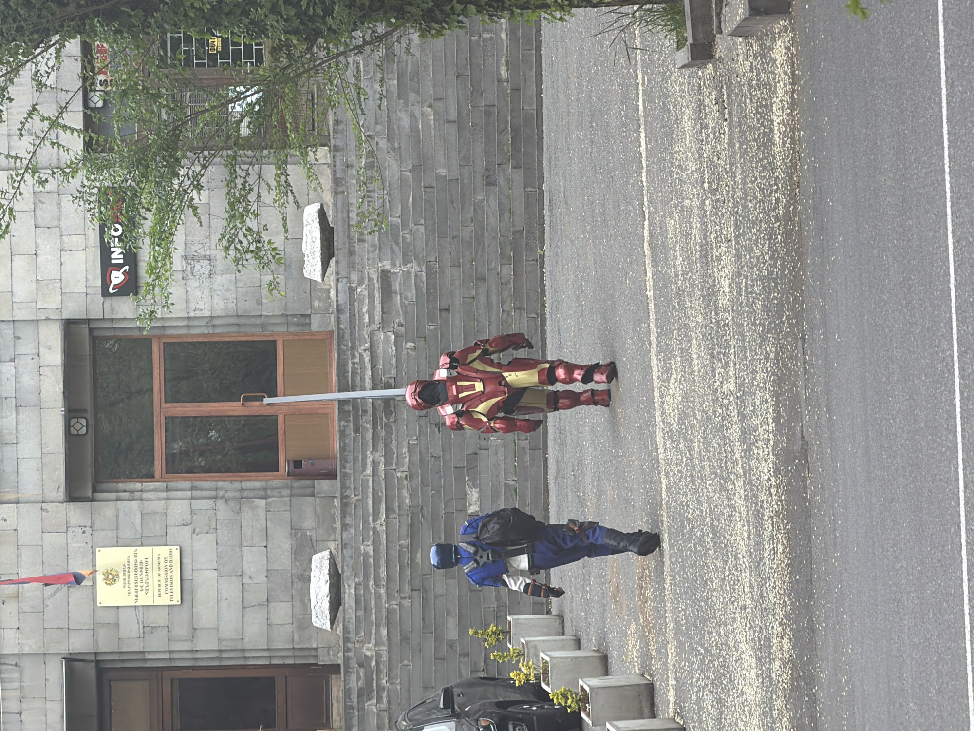 The image shows two individuals dressed in superhero costumes standing on a paved surface next to a building. The first figure is wearing a red and gold suit, resembling Iron Man, while the second is in a blue and gray outfit with a helmet. A black car is parked nearby. The building has a stone facade with windows and signage, including a plaque with Armenian text. A sign with "INFO" in large red letters with a black design is visible on the upper left side of the building. The architecture is characteristic of Yerevan, Armenia.