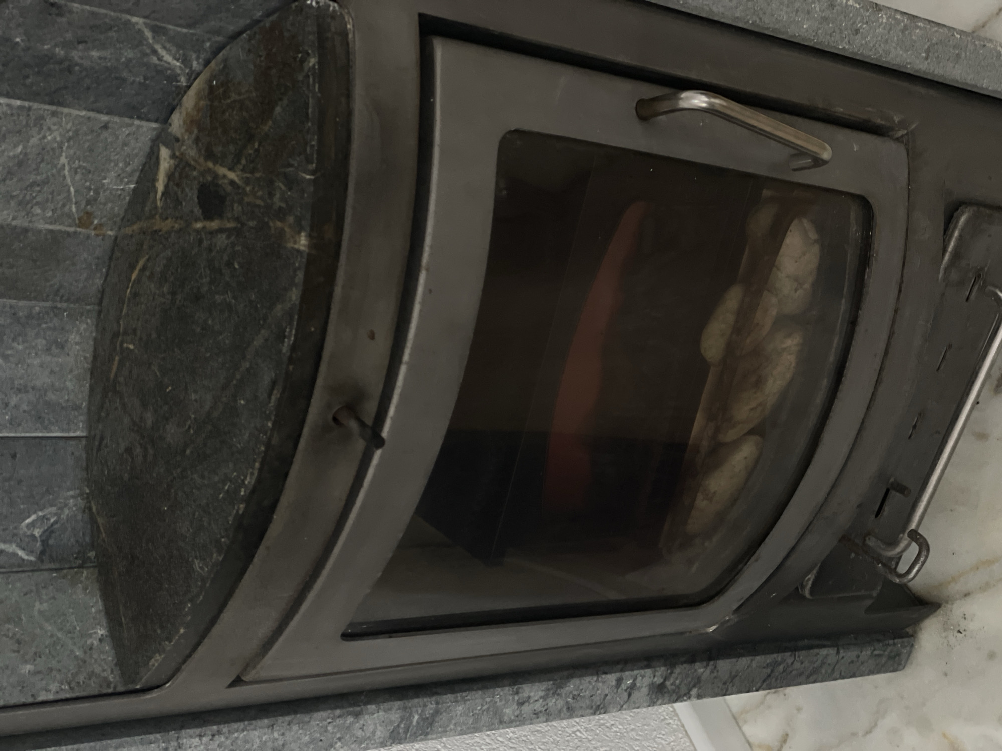 The image shows a close-up view of a dark grey marble-clad wood-burning stove. The stove has a curved glass door, through which several logs are visible inside. A metal handle is attached to the right side of the door. The stove is situated in a room with marble flooring and a textured wall to the left. The scene is well-lit. The specific location in Ulmen, Germany, is not determinable from the image alone.