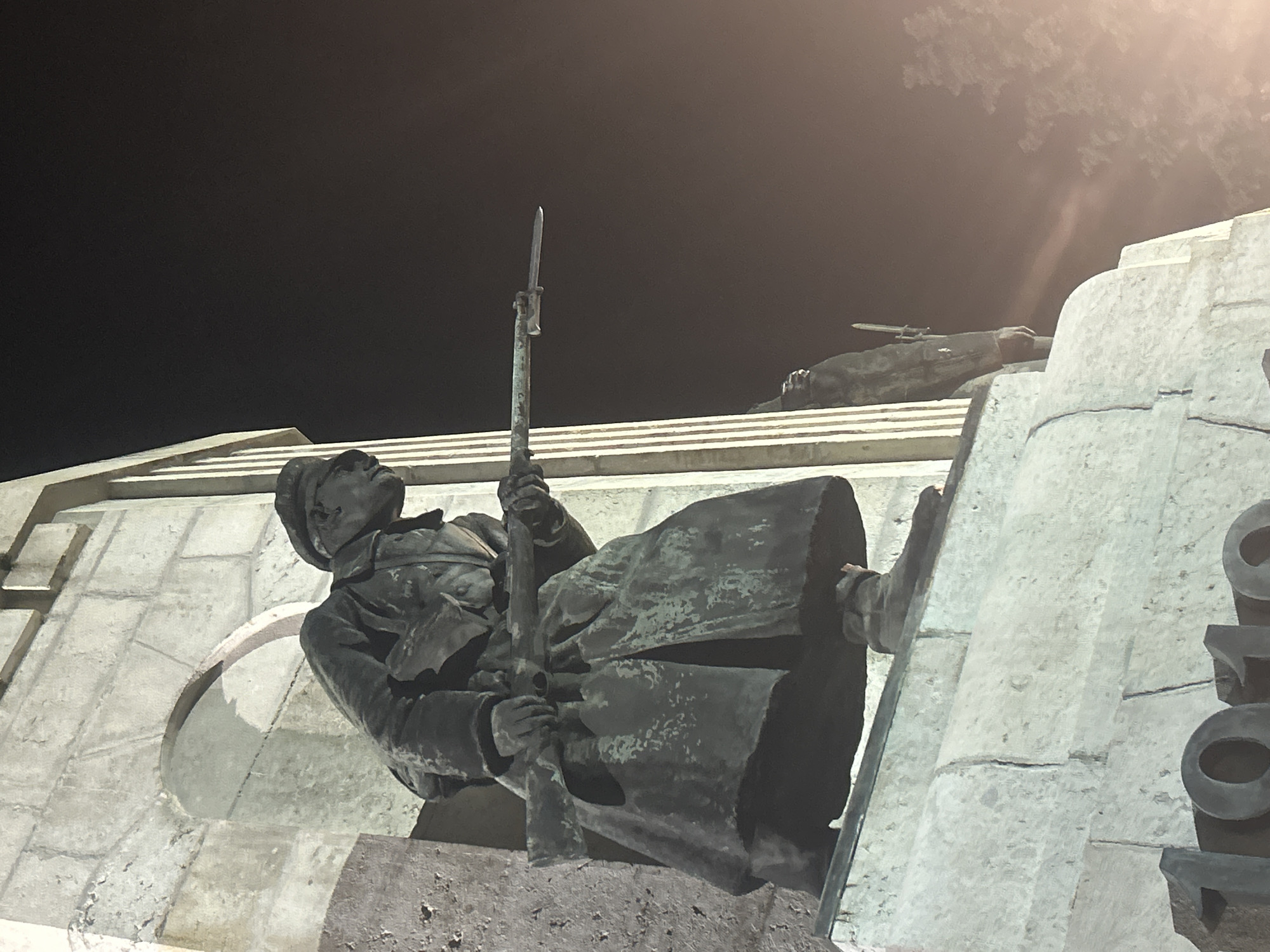 The image shows a monument in Veliko Tarnovo, Bulgaria, at night. The monument features bronze statues of soldiers. One statue depicts a soldier in uniform, holding a rifle with a bayonet fixed. Another statue shows a fallen soldier in the background. The statues are mounted on a stone structure. The background is dark, with a streetlight visible at the top right.