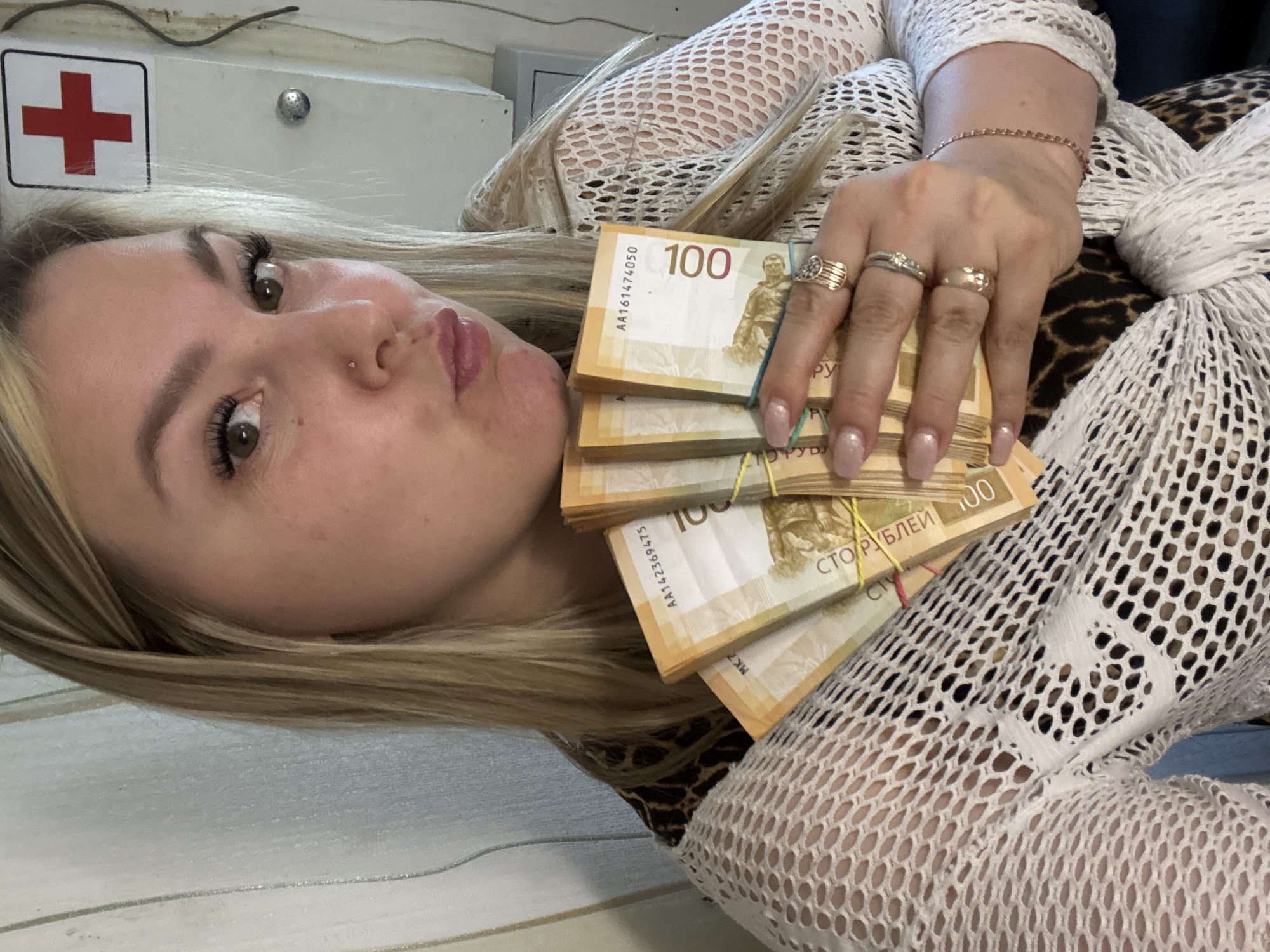 A woman with blonde hair and multiple rings on her fingers is holding several stacks of 100-ruble banknotes, secured with rubber bands. She is wearing a white fishnet-style top over a leopard-print garment. The background is a white surface with a medical sign featuring a red cross. The banknotes have "СТО РУБЛЕЙ" written on them. The location is not identifiable as Noviy, Russia, from the given image.