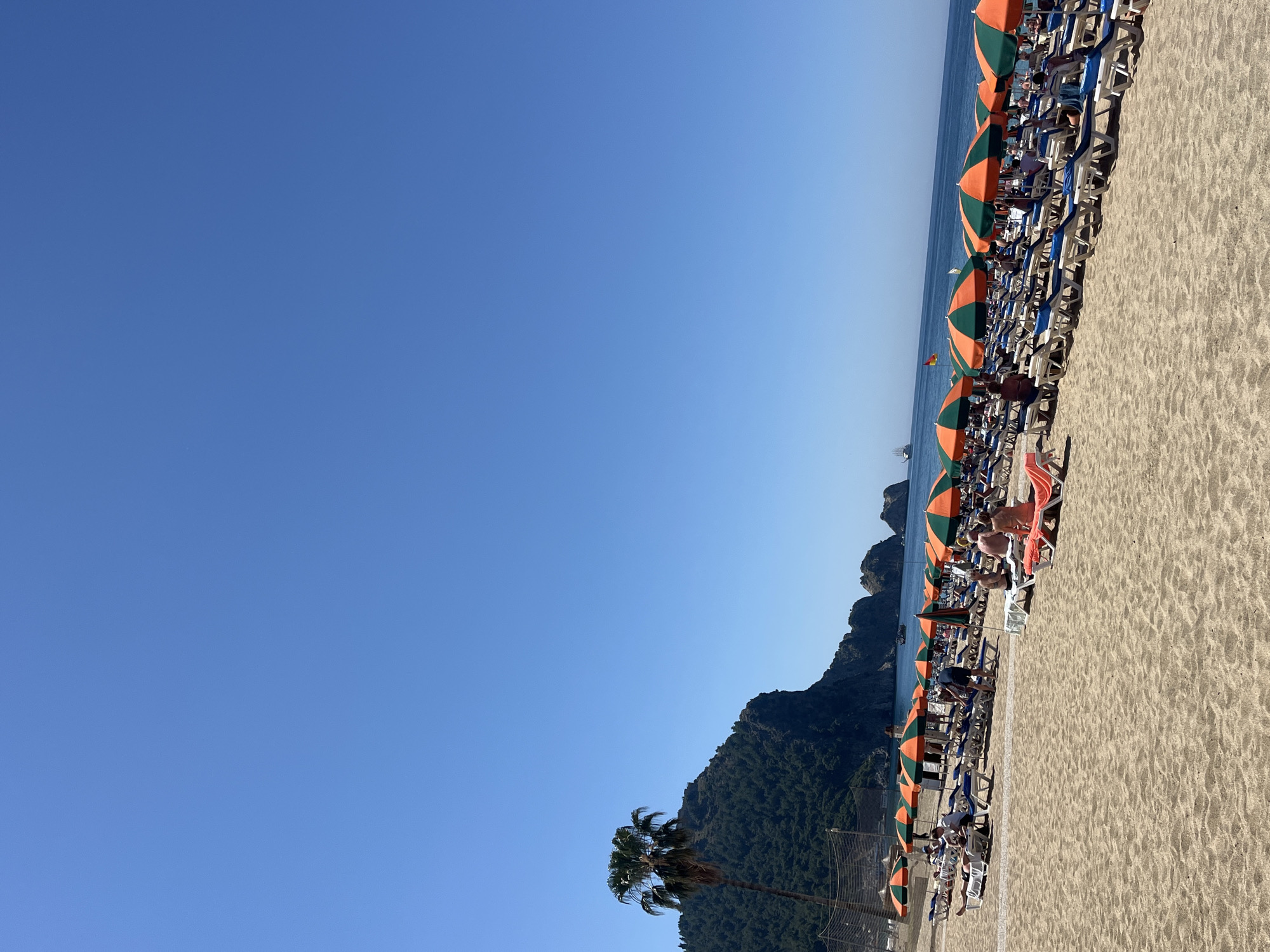 The image shows a beach in Alanya, Turkey. The beach is filled with orange and green striped umbrellas and beach chairs. People are lounging on the chairs. Behind the beach is a mountain covered in trees. The sky is blue.