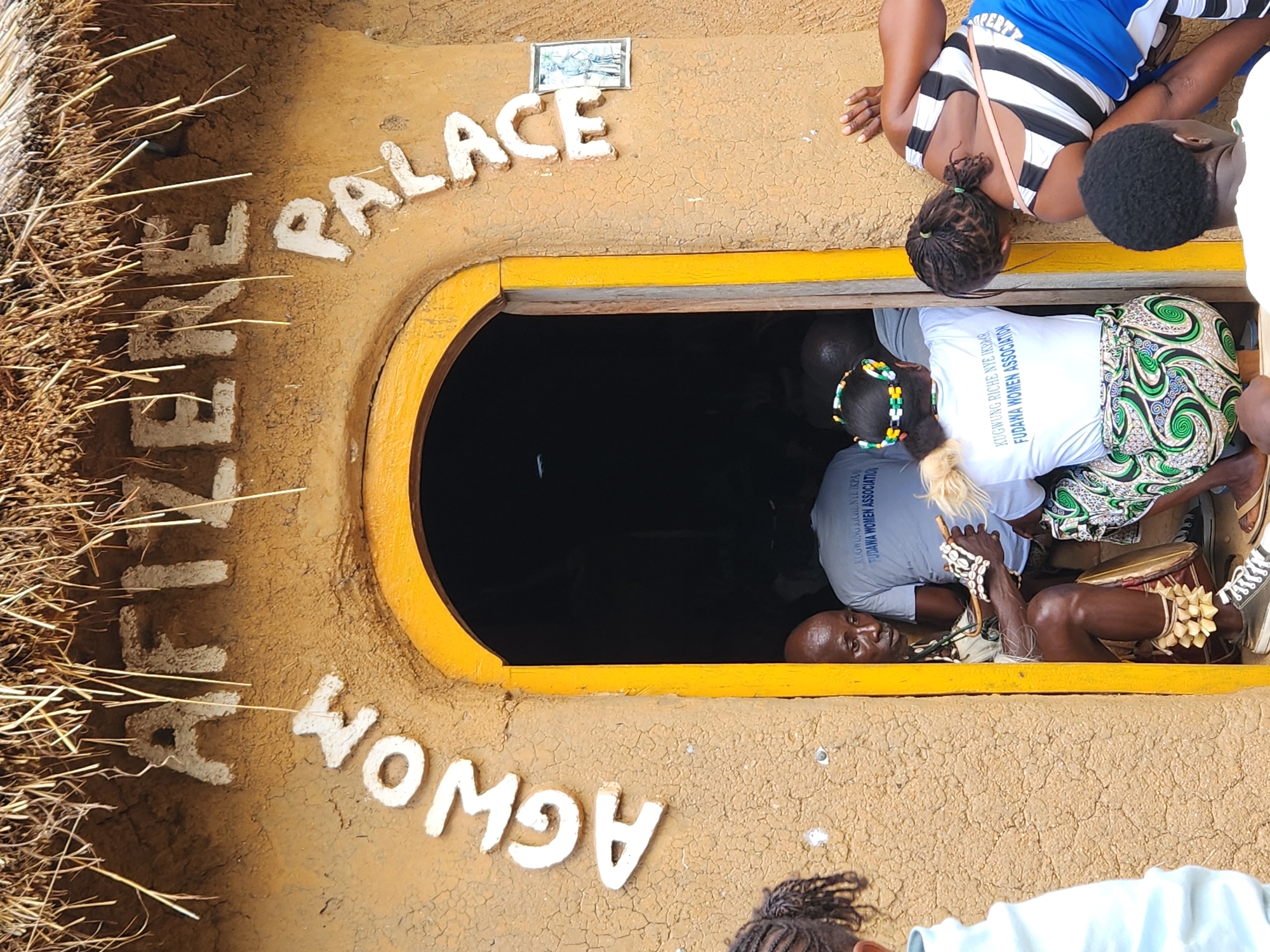 The image depicts an outdoor scene, likely in Jos, Nigeria, featuring an entrance to a building with text inscribed above it. The building's exterior is made of textured, earth-toned material, possibly adobe or clay, with a thatched roof visible on the left. White, raised letters spell out "YAFTZEREN PALACE" arching over the entrance, with "AGWOM" written below. The entrance itself is framed by a bright yellow border and opens into a dark interior.
Several people are visible in and around the entrance. In the foreground, a person's legs and feet are visible at the bottom right, wearing sneakers. Above the entrance, the upper bodies of several individuals are visible. One person wears a blue and white striped top, another a black top with a white stripe, and a third has dark curly hair. Inside the dark opening, a man in a grey t-shirt with text reads "KUGWUNG RICHE NYE IKPRAR FUDAWA WOMEN ASSOCIATION" is lying down. Another person in the entrance is wearing a white t-shirt with similar text. A woman inside the entrance wears a vibrant green and white patterned fabric. A drum and decorative items, including what appear to be shells and beads, are also visible. The lighting suggests it is daytime, and the overall atmosphere appears to be that of a gathering or event. The text indicates the presence of a "Fudawa Women Association".
