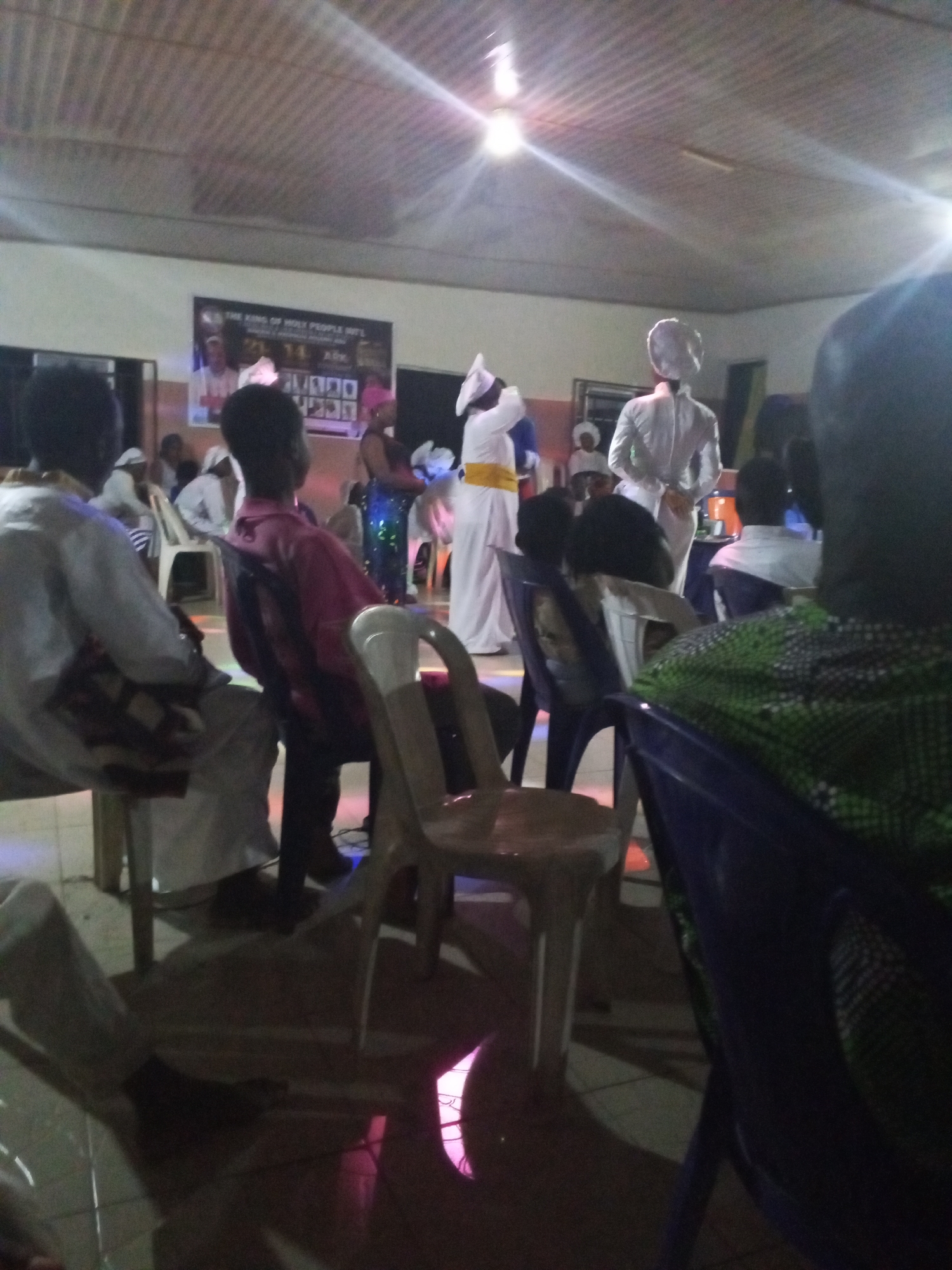 The image shows an indoor gathering in Lokoja, Nigeria. The room has a low ceiling with exposed beams and bright overhead lighting. A poster or banner is mounted on the wall towards the front of the room. Several people are seated on plastic chairs, facing the front. Some are dressed in white robes with head coverings, and at least one individual on stage is wearing a white robe with a yellow sash. Other attendees are wearing casual clothing. The flooring is tiled. A person in the foreground is partially visible, wearing a green patterned shirt.