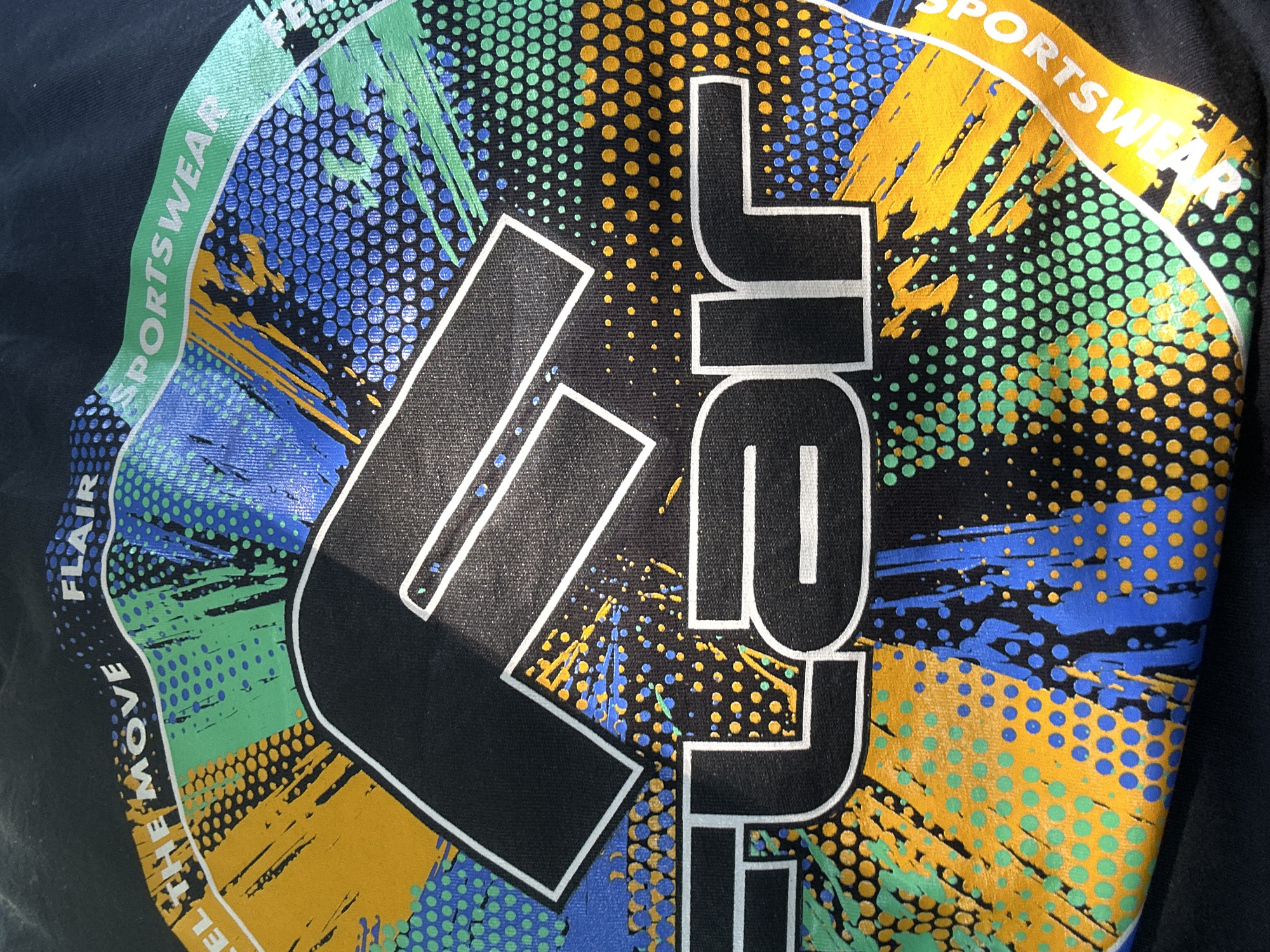The image shows a close-up of a black t-shirt with a colorful graphic design. The design features the word "FLAIR" prominently in large white-outlined black letters, with a stylized "F" above it. Surrounding the text is a dynamic pattern of abstract shapes, splatters, and dots in shades of yellow, green, blue, and white. There are also words "FEEL THE MOVE" and "SPORTSWEAR" integrated into the design on a curved band. The fabric of the t-shirt is visible, and it appears to be slightly creased.
This image is not taken in Plovdiv, Bulgaria. The provided context about the location is incorrect.