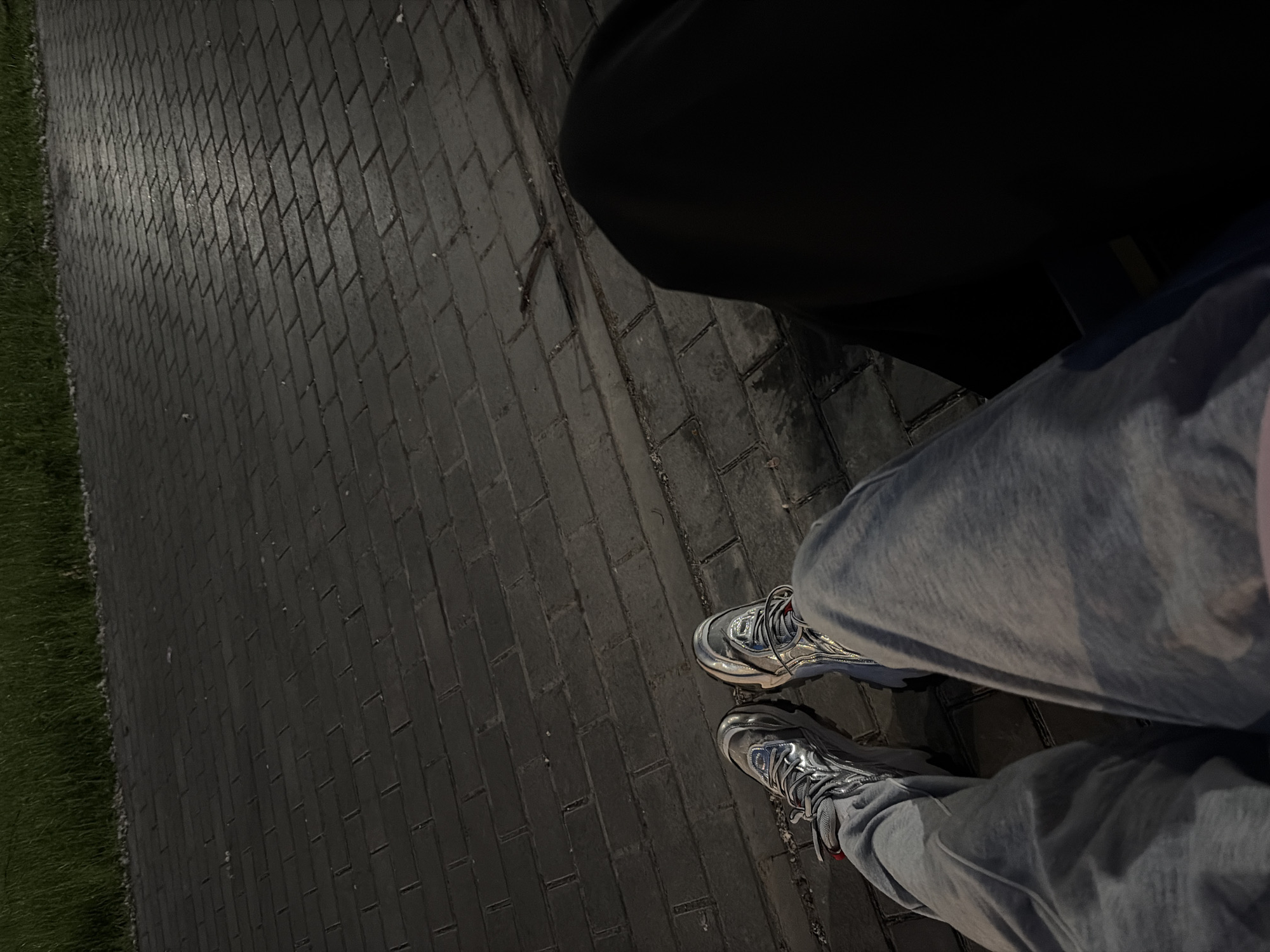 The image shows a close-up, high-angle view of two people's legs and feet sitting on what appears to be a paved surface, possibly a park bench. The person on the left is wearing grey sweatpants and metallic silver sneakers with red accents. The person on the right is mostly obscured by dark clothing, with only a portion of their leg visible. The ground is paved with dark rectangular bricks, and a strip of green grass is visible in the background. The lighting suggests it is either dusk or dawn, with a faint reflection on the bricks. The location is identified as Unknown, Unknown.