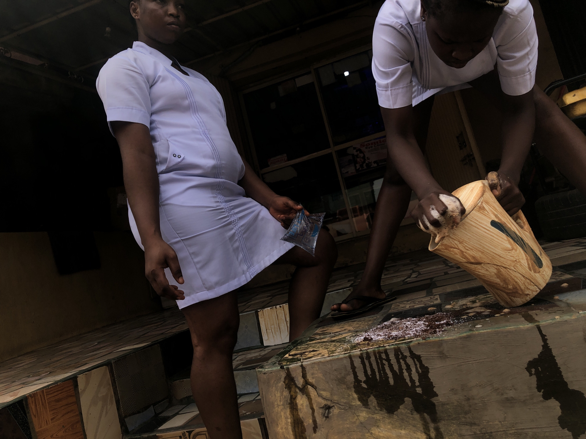 Two women are in the city of Obosi, Nigeria. The woman on the left is standing, wearing a white uniform dress with short sleeves and holding a small plastic bag with blue contents. The woman on the right is bending down and appears to be cleaning or scrubbing a light brown pitcher with soap. The area around the pitcher is wet and there are suds present. Both women are dark-skinned. The scene is set on a tiled surface. The lighting is dim.