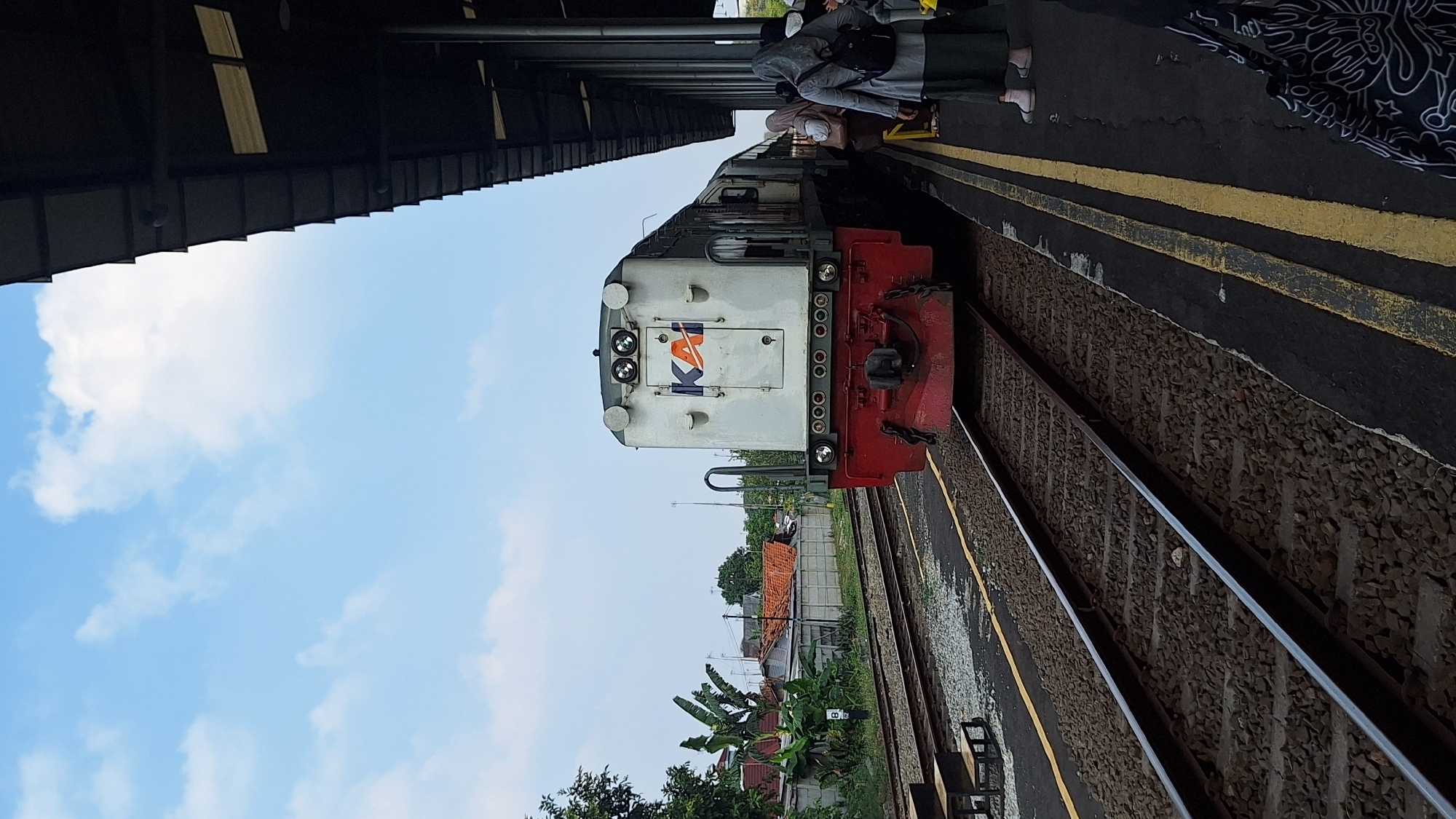 The image depicts a train at a station, possibly in Karawang, Indonesia. A white and red locomotive with the "KAI" logo is positioned on the tracks, facing the viewer. The train is at a platform where several individuals, dressed in modest attire, are waiting. One person, wearing a gray hijab and backpack, stands out. The station features a covered walkway with visible support pillars and a dark roof. The sky is clear with scattered clouds, and in the background, a residential area with houses and vegetation is visible. The ground is covered with gravel and ballast, typical of railway infrastructure. The scene appears to be in the daytime under natural light.