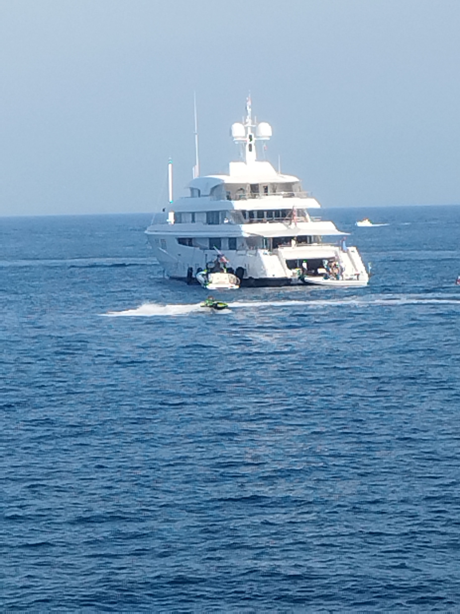 A large, white, multi-decked yacht is anchored in the blue waters off the coast of Antibes, France. The yacht has several levels and a tall mast. The stern of the yacht is open, revealing people on board. A smaller, white boat is docked at the stern. In the foreground, a green jet ski is being ridden by a person, leaving a trail of white spray on the water. Another smaller, white boat is visible in the distance to the right of the yacht. The sky is a clear, pale blue. The water is calm with gentle ripples. The scene suggests a sunny day with recreational activity on the water.