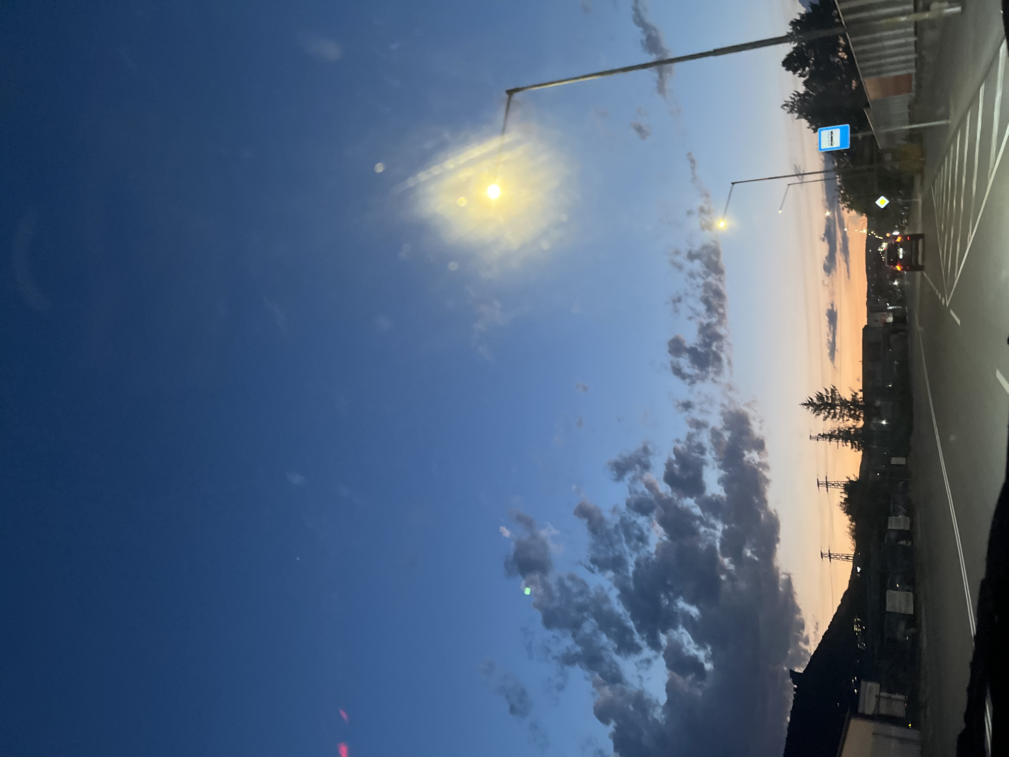 The image depicts a view from a car driving on a road at dusk, likely in the city of Shumen, Bulgaria. The sky is a deep blue with scattered clouds, transitioning to an orange and pink horizon. A street lamp is visible, illuminated and casting a bright light that creates a lens flare effect in the image. Further down the road, another street light is visible, and a blue bus stop sign is posted. The road has white lane markings. On the left side of the road, there are buildings and what appear to be power lines. On the right, there is a fence and some trees. A dark-colored car is in front of the camera. The overall scene suggests an urban or suburban setting during the transition from day to night.