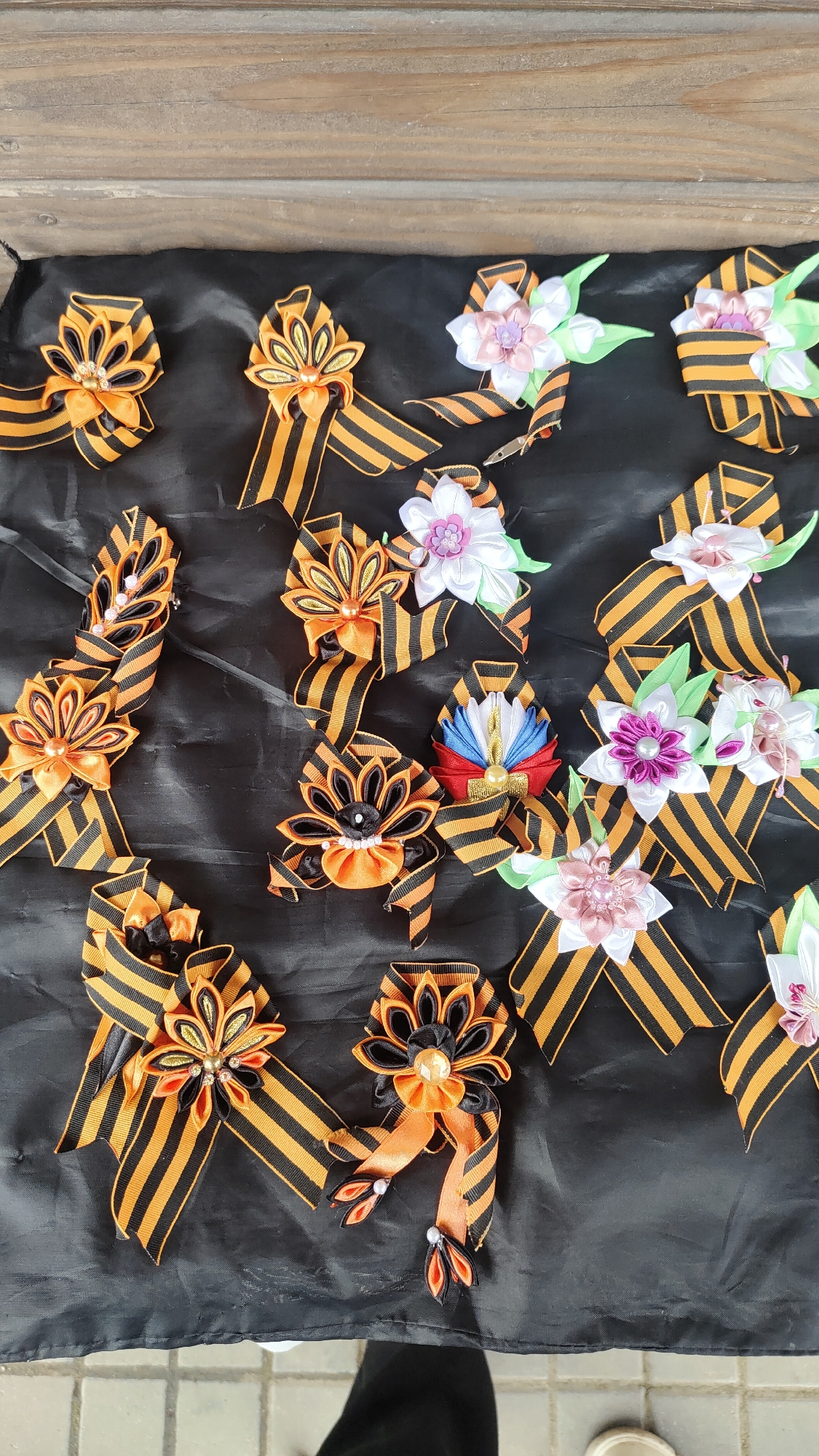 The image shows a collection of decorative ribbons and flower embellishments arranged on a dark fabric surface. The ribbons feature black and orange stripes. Some are adorned with fabric flowers in shades of white, pink, and purple, with green leaves. One ribbon features red, white, and blue elements. The scene is located outdoors with a glimpse of wooden surface at the top of the frame. The context and association of black and orange striped ribbons suggest it is related to Victory Day commemoration. While the location is not specifically indicated, it is likely in Krasnoyarsk, Russia, based on the prompt.