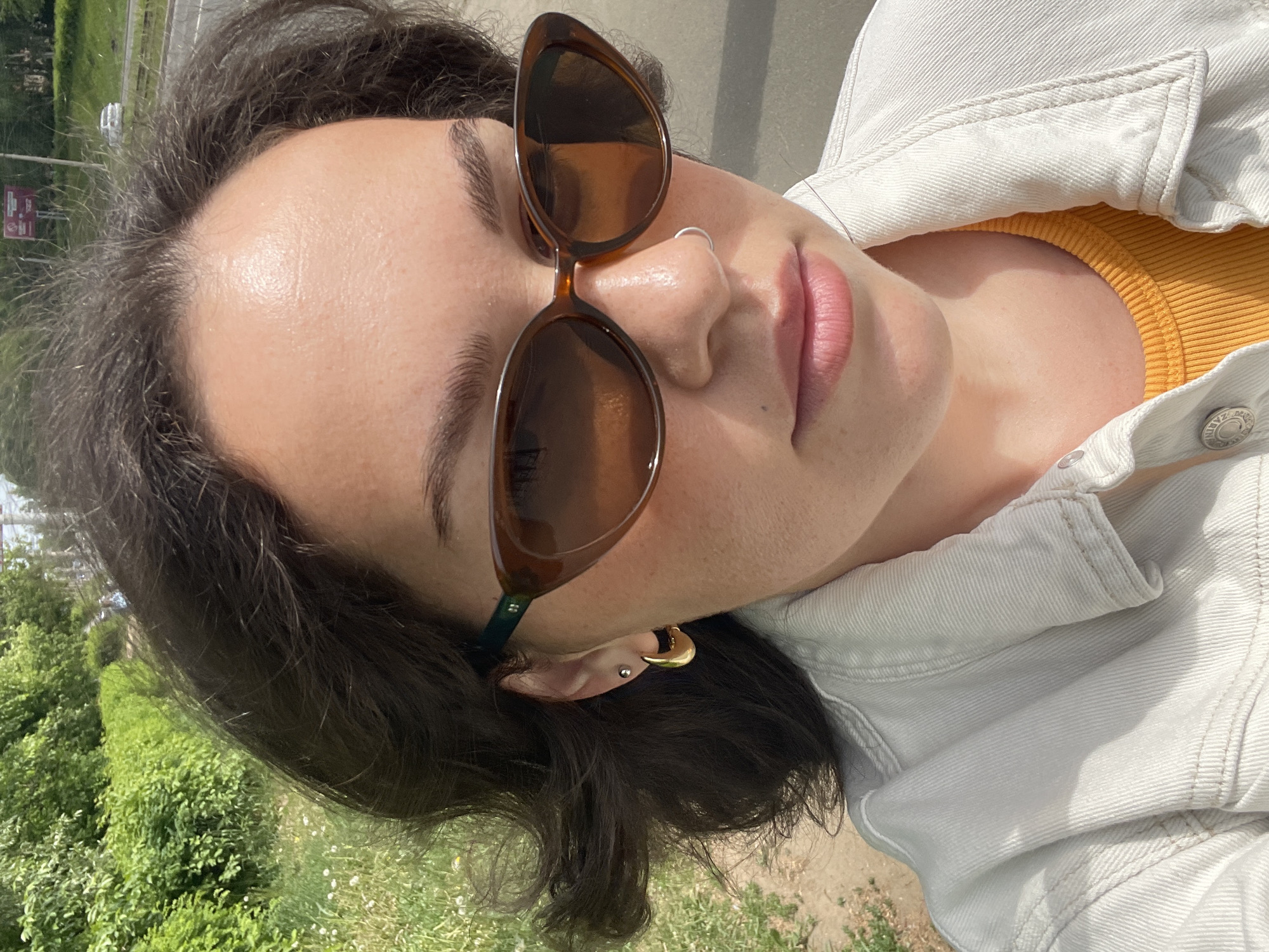 A woman is centered, facing the camera, wearing brown sunglasses and a white jean jacket over a yellow top. Her dark hair is visible. A nose ring is present. She has multiple ear piercings, including gold hoop earrings. She is outdoors, with greenery visible in the background. The location is not discernible from the image.