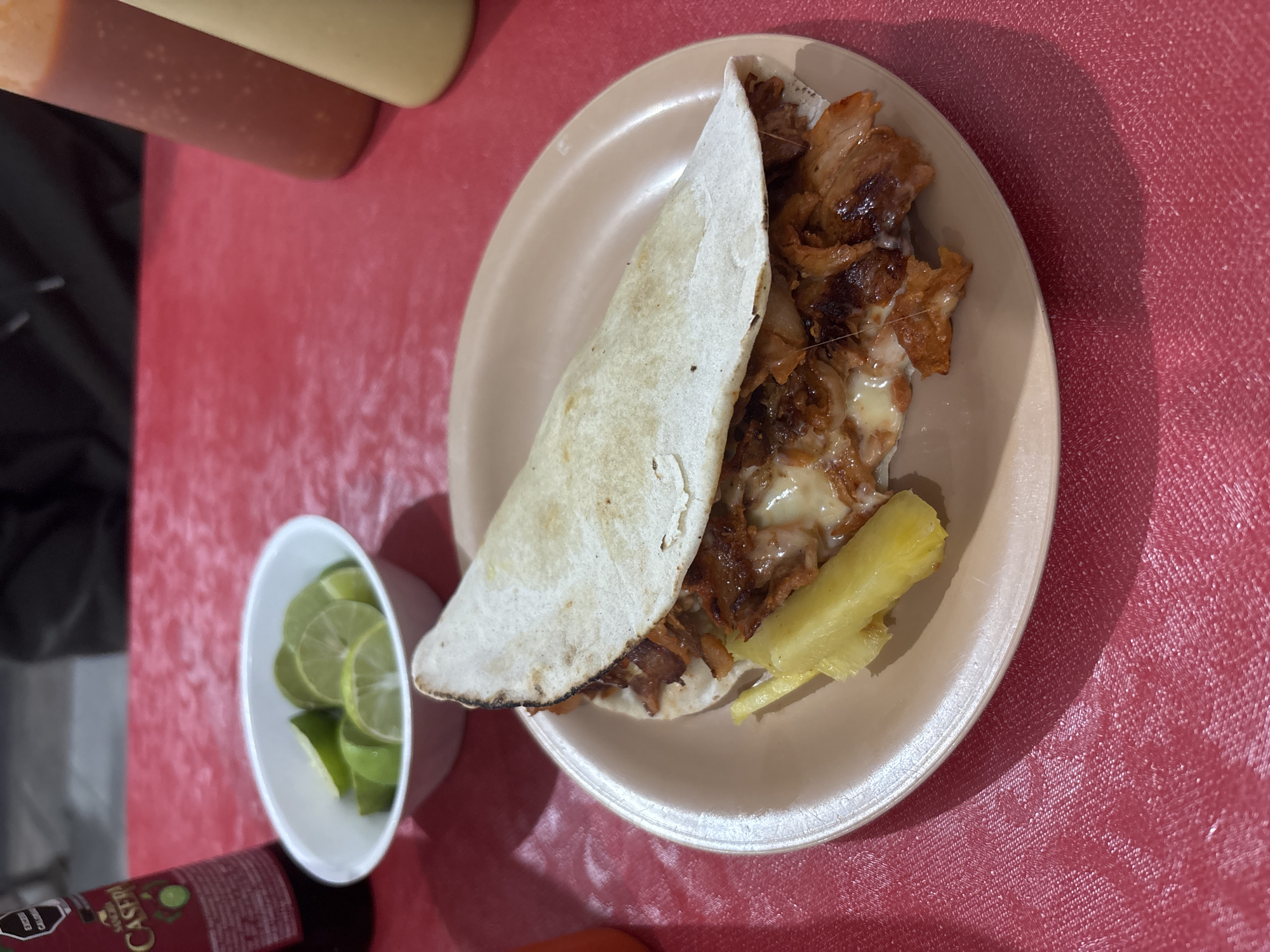 The image shows a taco al pastor on a beige plate. The taco is filled with marinated pork, melted cheese, and pineapple. A small white bowl filled with lime wedges is positioned to the left of the plate. Bottles of various sauces are visible in the upper left corner. The scene is set on a red tablecloth with a textured pattern. The image was taken in San Andrés Cholula, Mexico. A bottle of Casera apple soda sits at the bottom left.