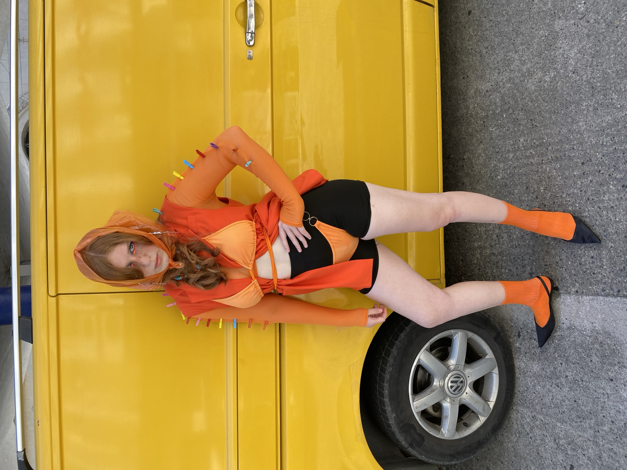 A young person with red hair is standing in front of a yellow van in Sochi, Russia. They are wearing an orange headscarf, an orange bikini top, an orange mesh shirt with clothespins attached, black shorts, orange socks, and black pointed shoes. The person is posing with one hand on their hip. The van is parked on a grey asphalt surface.