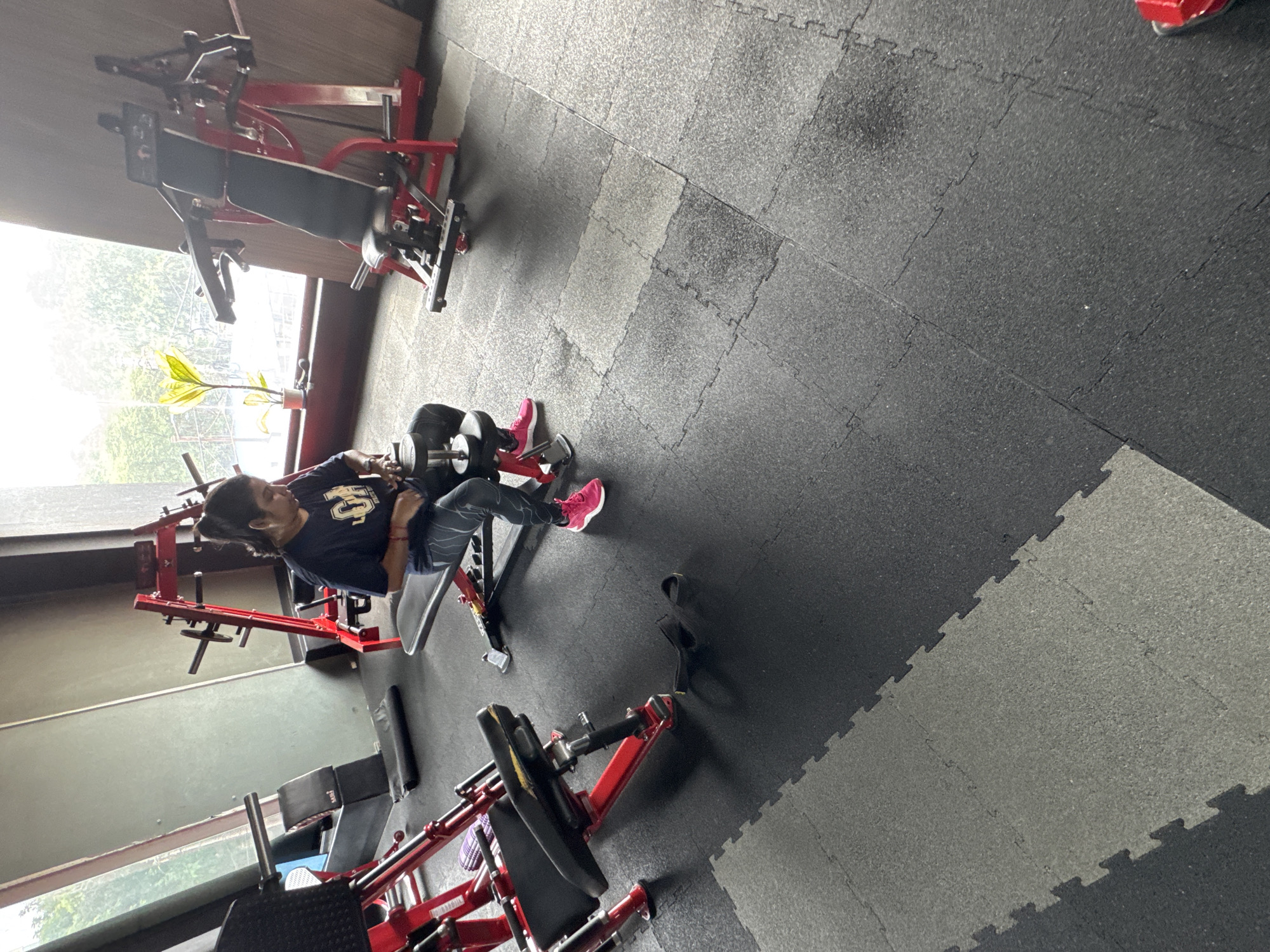 A woman is in a gym, sitting on a bench with a dumbbell in her hands. She is wearing a dark blue t-shirt and black leggings with a white pattern. Her hair is pulled back in a ponytail. The gym has red and black equipment, including weight machines and a bench. The floor is covered with black and grey interlocking mats. The background shows a large window with greenery outside. The lighting suggests an indoor setting, possibly during daytime. The scene appears to be a workout session in a gym. Based on the presence of specific gym equipment and overall ambiance, this image is likely taken in a gym located in Kolkata, India.