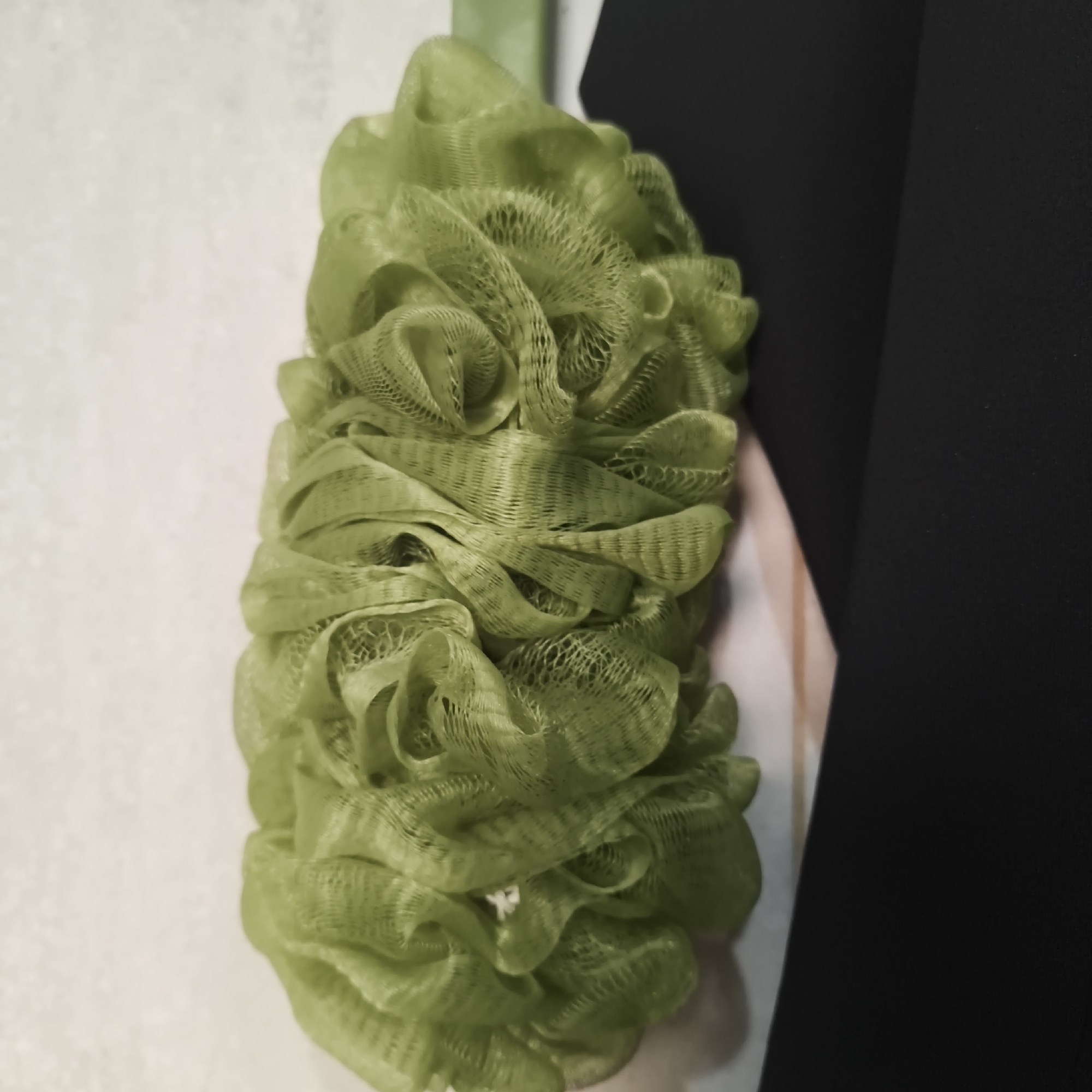 The image shows a green loofah hanging against a white textured wall, partially obscured by a black fabric. There are no people or identifiable landmarks present, and therefore its location in the city of South Jakarta, Indonesia cannot be determined.