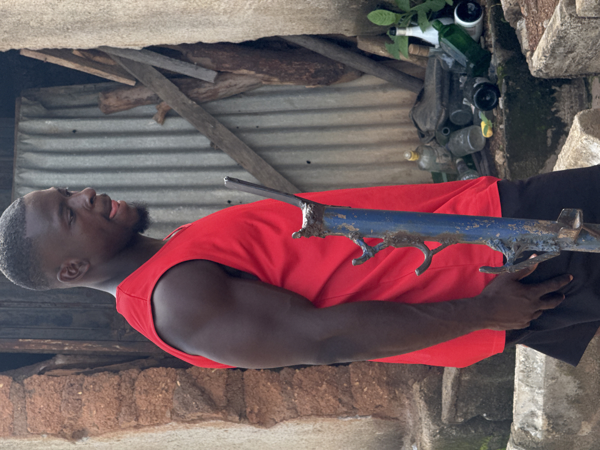 A dark-skinned man in a red tank top and black shorts stands in profile, facing right. He has a short haircut and a beard. His right arm is bent, with his hand resting on his shorts. He appears to be walking. The background shows a corrugated metal wall, some wooden planks, and a pile of discarded bottles, including clear glass, green glass, and a white-capped bottle. There are also some plants and a rough stone structure. The scene suggests an outdoor environment, possibly an alley or a neglected area within Akwanga, Nigeria.