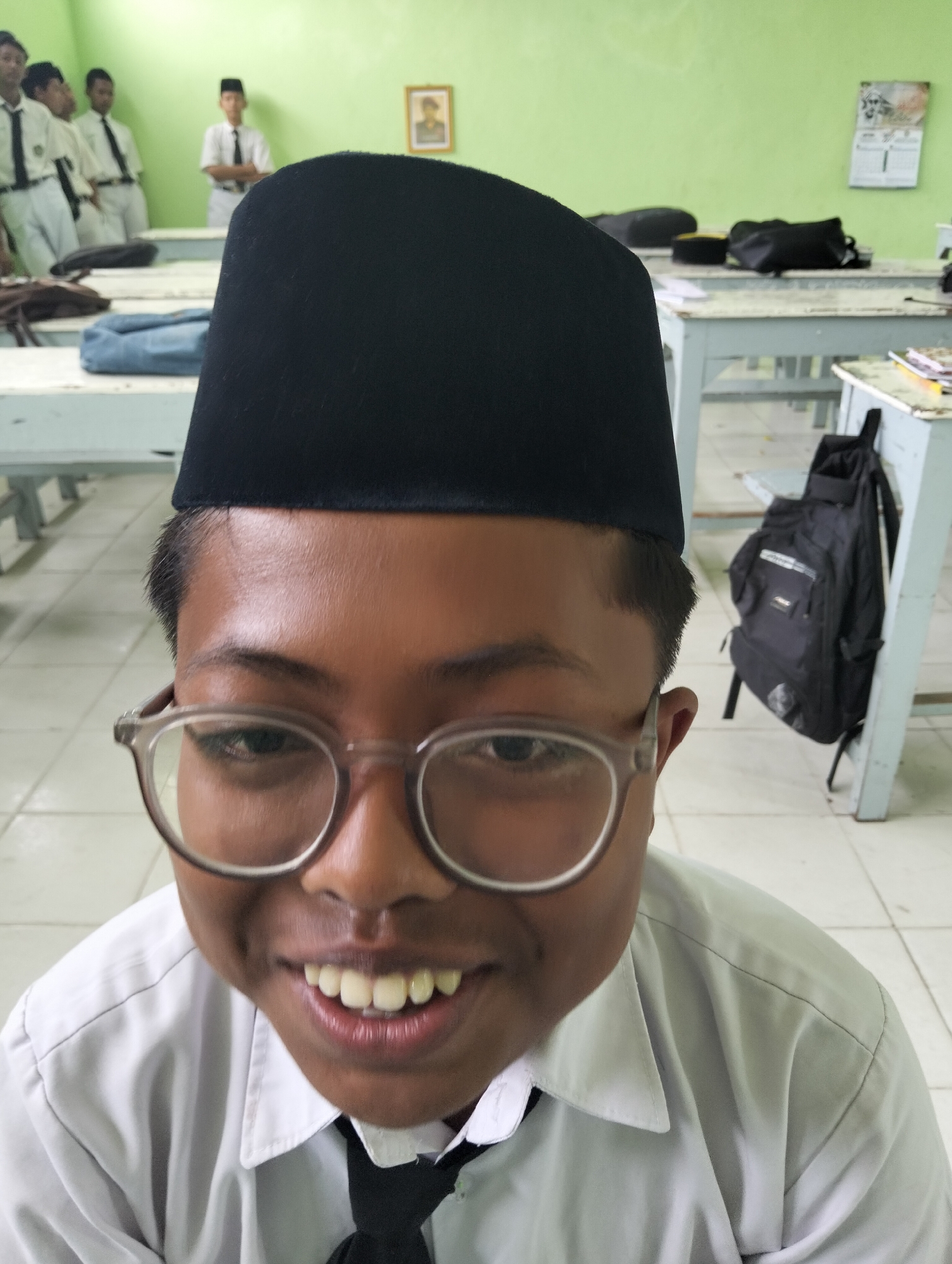 The image shows a close-up of a young boy wearing a black peci (a cap) and glasses, smiling. He is in a classroom setting, as evidenced by the desks and chairs visible in the background. Several other students are also present in the background, some of whom appear to be wearing school uniforms. The classroom walls are painted green, and there are pictures and a calendar hanging on the wall. A black backpack is visible on a chair. The image appears to have been taken in Kranji, Indonesia, as indicated by the presence of students in what is likely a school uniform and the cultural context of the peci. The overall atmosphere seems casual and relaxed.