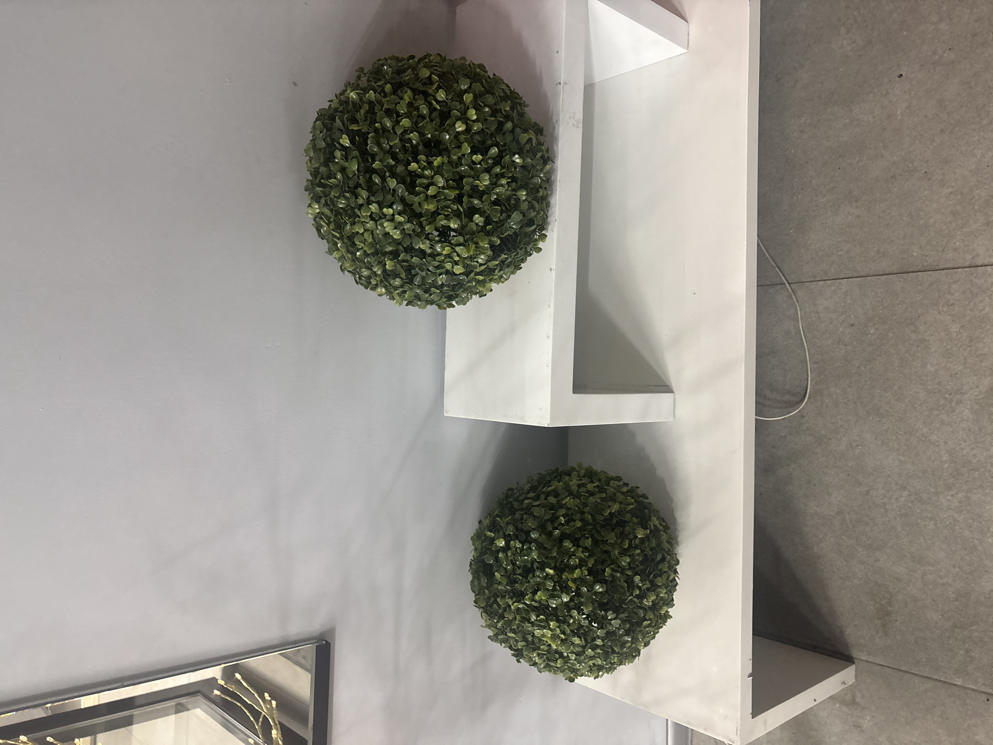 The image, taken in Solontsy, Russia, displays an indoor setting featuring decorative elements. Two spherical faux topiary plants with dense green leaves are prominent. One topiary is positioned higher up on a white shelving unit, while the other sits below it, resting against the same unit and the wall.
The shelving unit is white, appearing to be a modern, angular design, possibly a corner shelf, and is clean and minimalist. Behind the topiaries and the shelving unit is a light grey wall. To the right of the shelving unit, the floor is visible, covered in dark grey tiles with a visible grout line. A thin white electrical cord runs across the tiled floor.
In the bottom left corner of the image, partially visible, is a section of a framed mirror or artwork. Within the frame, a reflective surface shows a golden, abstract, branch-like design.
There are no people present, and no specific activity is taking place. The scene suggests a static, decorated interior, possibly a showroom, an office, or a modern living space. The lighting appears to be artificial, typical of an indoor environment, and no specific time of day or weather conditions can be inferred. There is no visible text in the image.