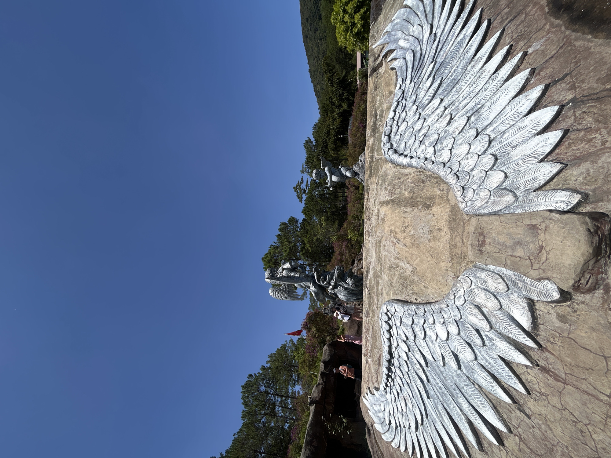 This image captures a vibrant outdoor scene at an attraction in Da Lat, Vietnam, under a clear, bright blue sky.
In the foreground, a prominent pair of large, silver-colored angel wings are intricately carved into a beige or reddish-brown rock formation. These wings are designed for visitors to stand between them for a photo opportunity. The texture of the rock surface is visible, showing some cracks and variations in color.
Beyond the foreground wings, the scene unfolds into a lush landscape dominated by dense green foliage and tall trees covering a hilly terrain. Scattered throughout this area are several artistic sculptures. On the left side, closer to the foreground, a smaller statue of a cherub or angel figure is perched on a rock. Further back, a larger, more elaborate statue depicting a mythical creature or guardian figure with a dynamic pose is visible amidst the trees.
Several people are dispersed throughout the mid-ground, walking along paths and observing the sculptures, suggesting a leisurely visit to a park or garden. A small red flag, possibly the national flag of Vietnam, is seen in the distance, adding a pop of color to the green and blue palette.
The bright daylight and absence of clouds indicate sunny weather, with strong shadows cast by the wings and other objects suggesting it is midday or early afternoon. The overall atmosphere is serene and picturesque, highlighting both natural beauty and human artistry. No discernible text is visible in the image.