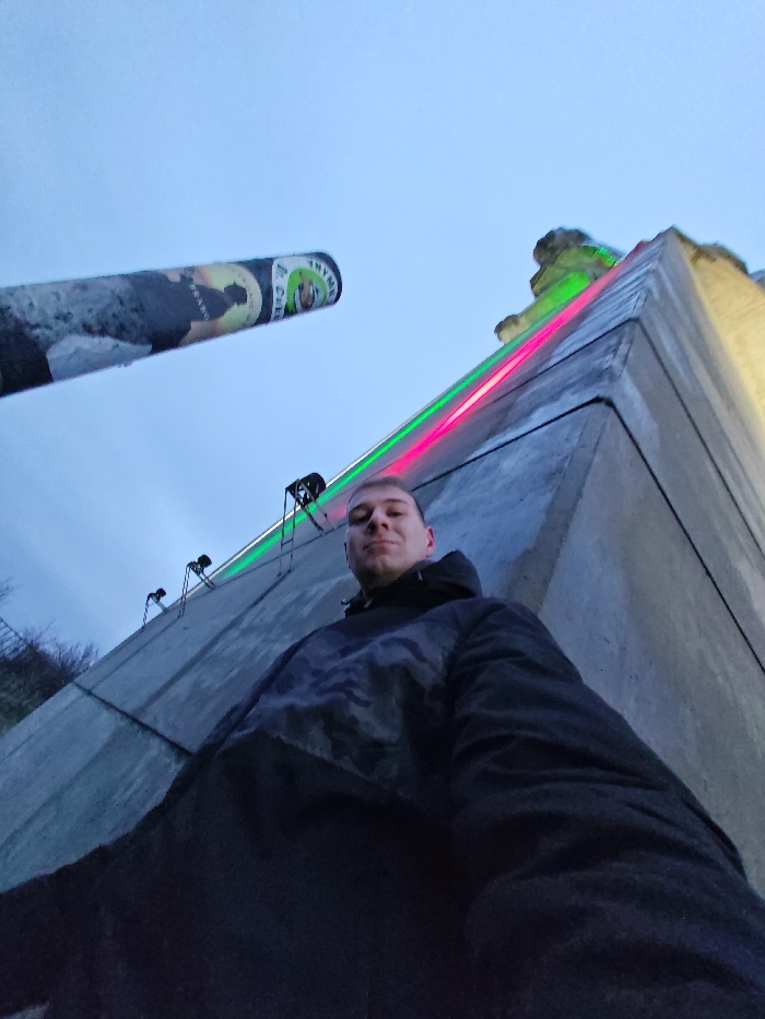 The image displays a low-angle perspective of a male individual positioned in the foreground, looking directly at the camera with a subtle smile. He is wearing a dark, hooded jacket with a grey patterned design across the chest.
Behind the individual, a massive, angular concrete structure ascends prominently, identifiable as part of the Monument to 1300 Years of Bulgaria (Founders Monument) in Shumen, Bulgaria. This structure features two parallel neon light strips—one green and one magenta—running vertically along a leading edge. Above these lights, several black spotlights are mounted. The uppermost visible section of the concrete structure displays an abstract architectural element.
Extending into the frame from the upper-left, a large, cylindrical object, resembling a cannon barrel, is visible. Its surface exhibits peeling material or graffiti, including a partial label with the text "J. COLE" and a green circular logo.
The sky in the upper portion of the image is a uniform light blue-grey, indicative of twilight or overcast conditions. The composition highlights the monumental scale of the background structures relative to the individual.