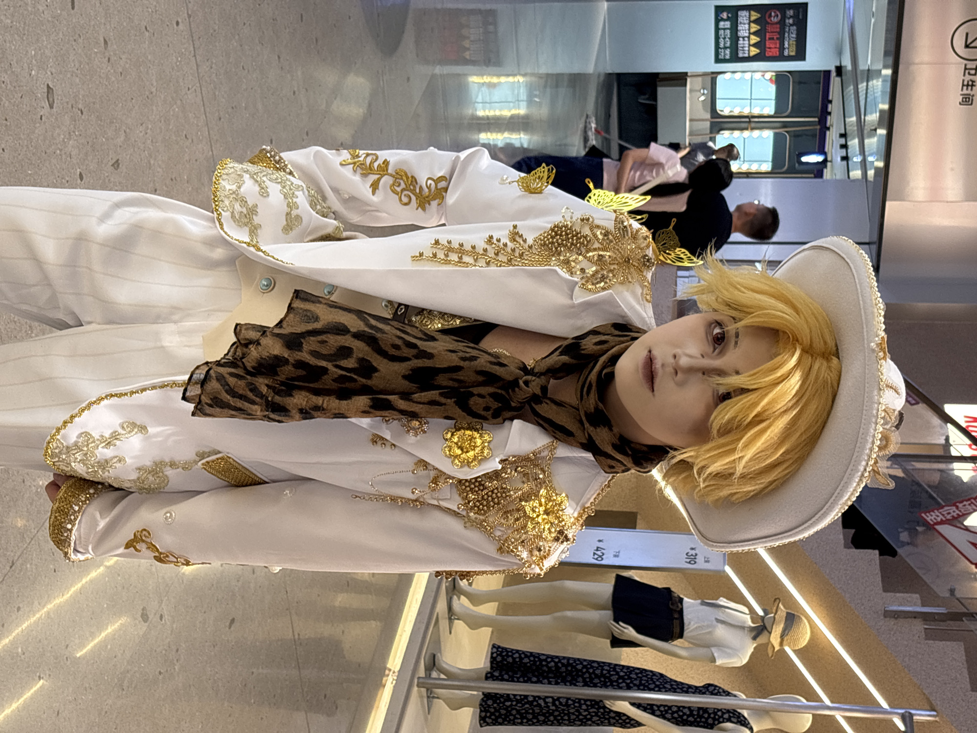 Here is a detailed description of the image:
**Scene:** An indoor setting, likely a shopping mall or retail space in Liangjiang, China.
**Subject:** A person dressed in a white and gold embellished suit with a leopard print scarf and a wide-brimmed hat. They appear to be cosplaying, possibly as a character with a flamboyant or aristocratic style. Their eyes have a reddish tint, likely due to makeup or editing.
**Surroundings:**
* Clothing display: Mannequins wearing dresses are visible in the foreground.
* Lighting: Bright fluorescent lights illuminate the area.
* Chinese signage: In the background, there's signage with Chinese characters, including "卫生间" (Wèishēngjiān), meaning "toilet" or "restroom," and various advertisement text.
* People: Blurred figures of other shoppers are seen in the background.
**Composition:** The photo appears to be taken at eye level. The subject is in the center of the frame, slightly tilted.
**Overall:** The image presents a scene with a person in distinctive attire within a commercial environment.