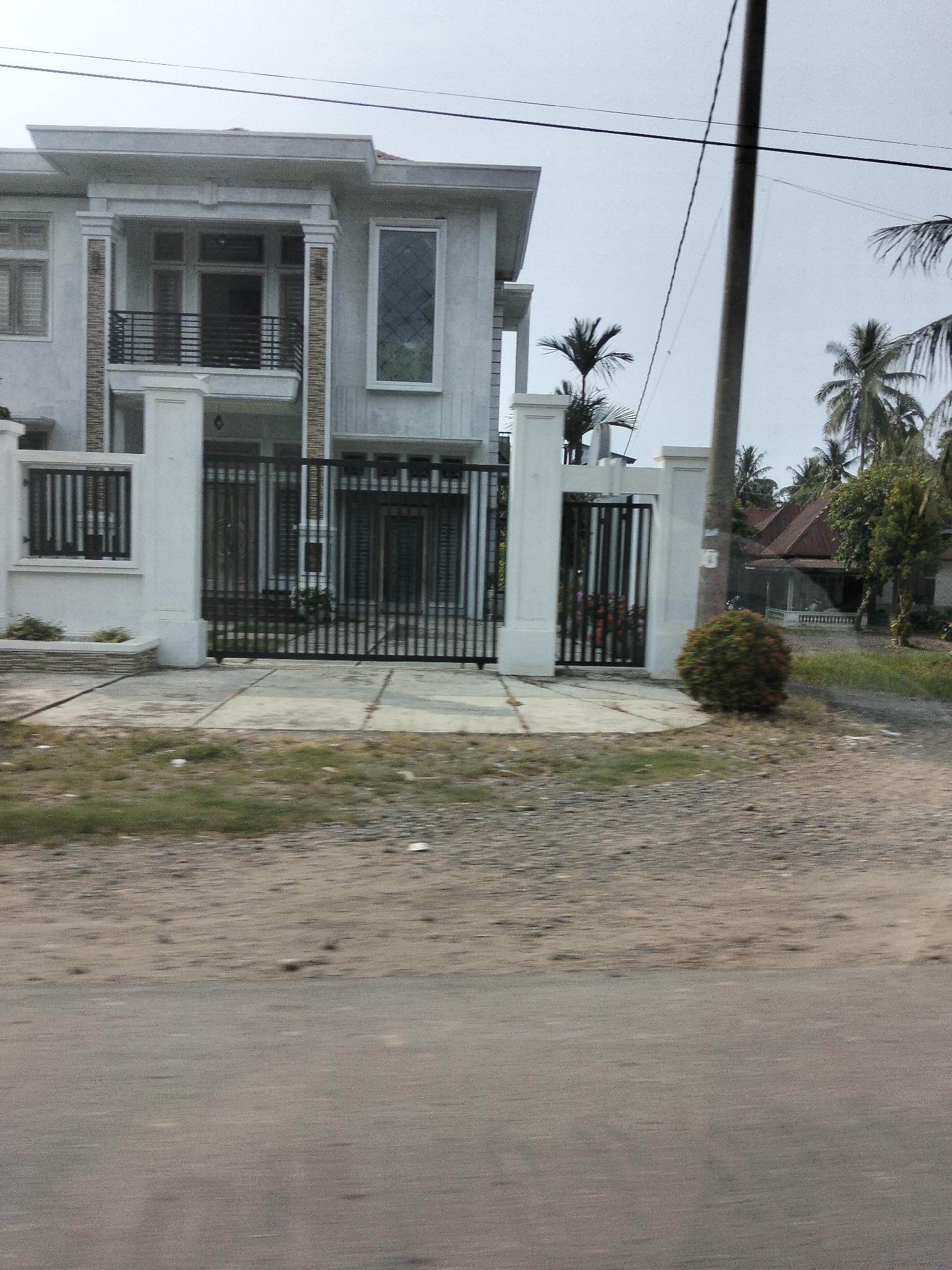 The image depicts a modern, two-story house with a white exterior, black metal gates, and a long driveway. The house features a balcony on the second floor with a black metal railing, a large window with a diamond pattern on the right side of the facade, and several other windows and doors. A black metal fence runs along the front of the property. Palm trees are visible in the background, along with a separate, older-style house with a reddish-brown roof. The scene is set on a clear day, with a cloudy sky. The foreground shows a rough, gravelly road. The location is identified as Tingkeummanyang, Indonesia. There are no people or animals visible in the image, nor are any actions or interactions taking place. The overall impression is one of a suburban or semi-rural residential area.