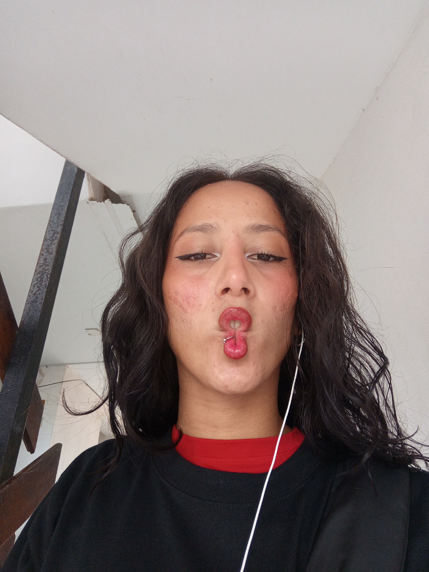 A young woman is posing for a selfie. She has dark wavy hair, fair skin, and is wearing a black long-sleeved shirt with a red collar. She is making a fish face with her lips and has a piercing in the middle of her bottom lip. She is wearing eyeliner and has white eyeliner on her lower lash line. There are acne scars on her cheeks. An earbud cord is hanging down from her ear. Behind her is a white wall and a metal railing. While it is impossible to confirm the location is Porlamar, Venezuela, based on the image alone, the architectural style and general appearance are consistent with urban areas in Venezuela.