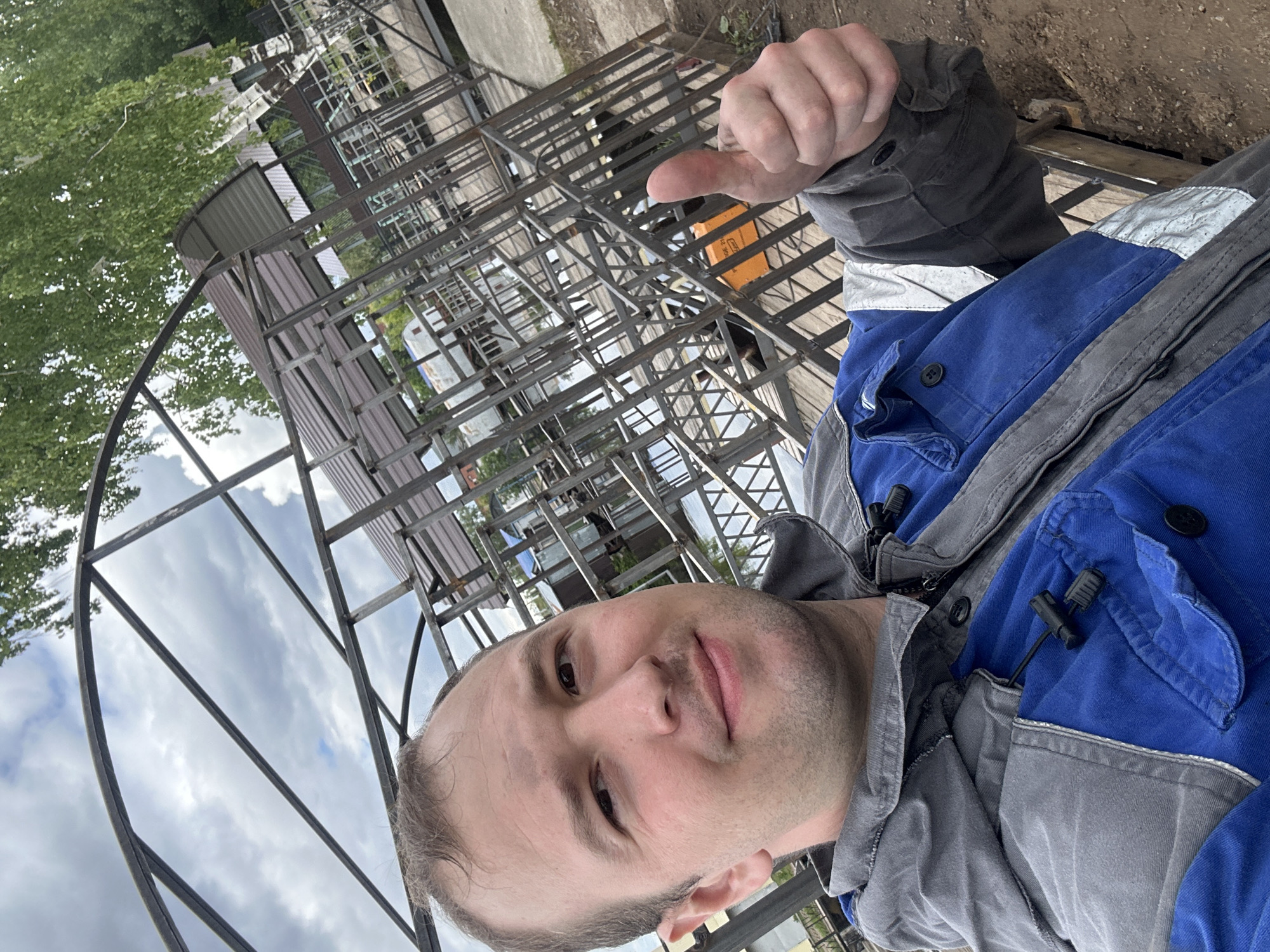 A man is captured in a selfie outdoors, giving a thumbs-up gesture, indicating approval or satisfaction. He is a Caucasian male with short light brown hair and a clean-shaven face, looking directly at the camera with a neutral to slightly smiling expression. He wears a two-tone work jacket: a blue torso with grey sleeves, collar, and side panels. Reflective silver strips are visible on the upper arms of the jacket. A small black clip, likely for a microphone or earpiece, is attached to the blue section of his jacket near the collar.
The scene behind him suggests an industrial or construction environment. Numerous intricate metal structures, resembling storage racks, shelving units, or framework for a building, fill the background. Some sections of corrugated metal, possibly roofing or siding, are visible within these structures. A bright orange object, possibly a box, can be seen among the metalwork. To the left, a large, curved metal archway is prominent, extending upwards towards a cloudy sky. Green foliage from trees is visible behind this archway and in the upper left of the frame. The ground appears to be unpaved, consisting of dirt or gravel. The overall impression is an outdoor workspace under an overcast sky during daytime in Chaikovsky, Russia.