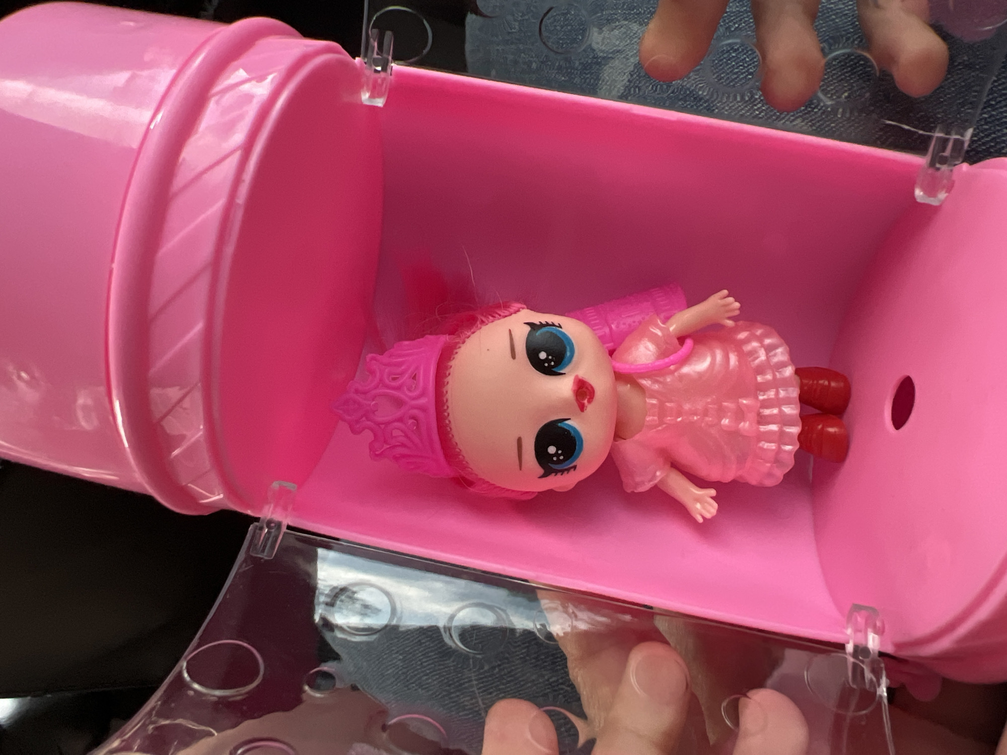 The image shows a pink LOL Surprise doll inside a pink plastic container. The container is partially open, with a clear plastic lid featuring circular indentations. The doll has pink hair, a pink dress, and red shoes. The container appears to be held by a person, whose fingers are visible at the top and bottom of the frame. The background is out of focus, showing hints of denim jeans and a dark surface. There is no information to indicate this photo was taken in Yekaterinburg, Russia.