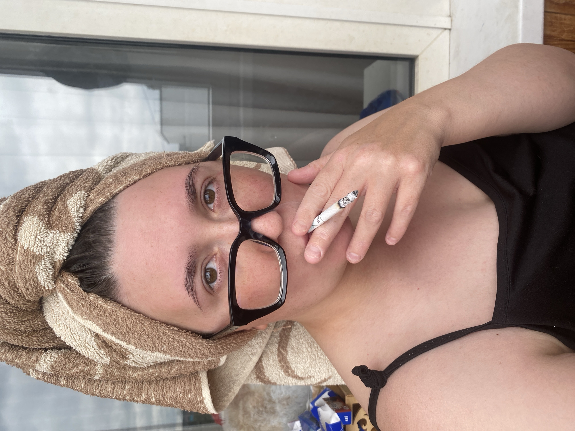 A woman with a towel wrapped around her head and black-rimmed glasses is holding a lit cigarette to her mouth. She is wearing a black tank top. She is indoors near a window with a view of what appears to be a city skyline. The image was taken in Chaikovsky, Russia.