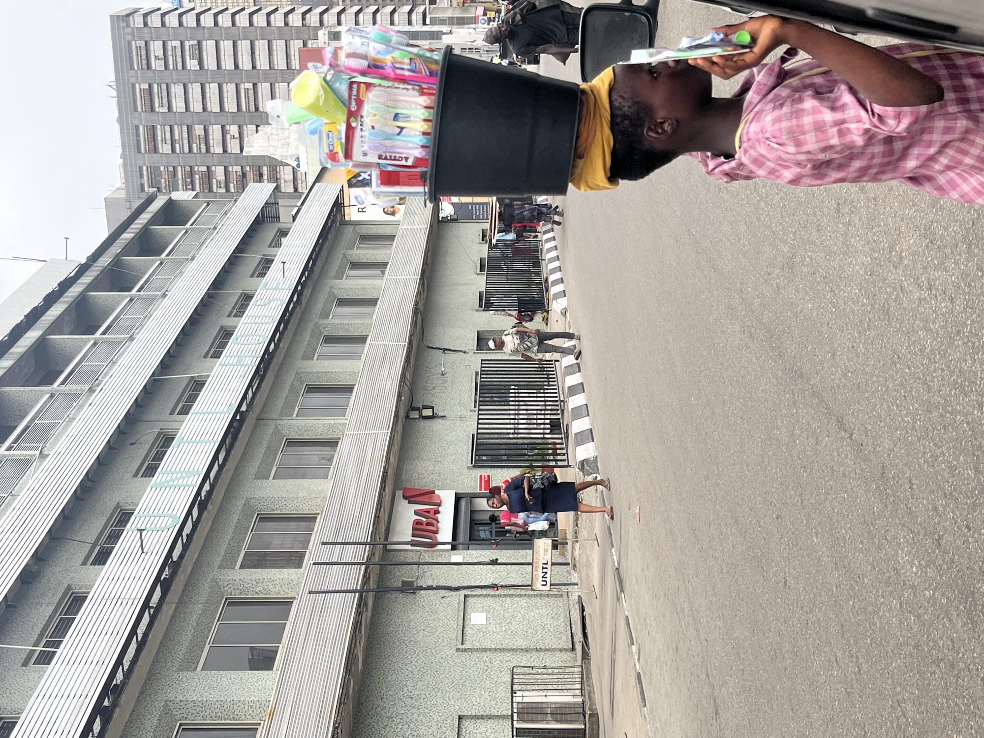 A street vendor in Lagos, Nigeria is balancing a large bucket of toothbrushes and other dental products on their head. The vendor is wearing a pink plaid shirt and a yellow head covering. The vendor appears to be holding a toothbrush to their face. The vendor is standing on a city street. A tall building is behind the vendor with a sign for "UBAN" on the front. Cars and people are moving around on the street. The sky is overcast.